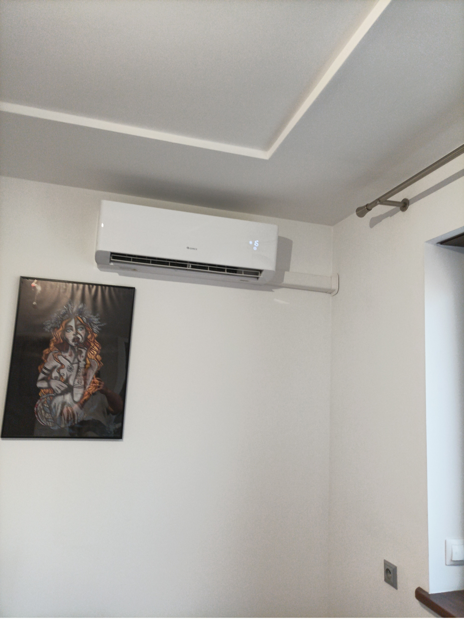 Biała klimatyzacja na ścianie w pokoju, obok wisi obraz w czarnej ramie. Widoczny fragment sufitu z ozdobnym wgłębieniem i karnisz nad oknem.