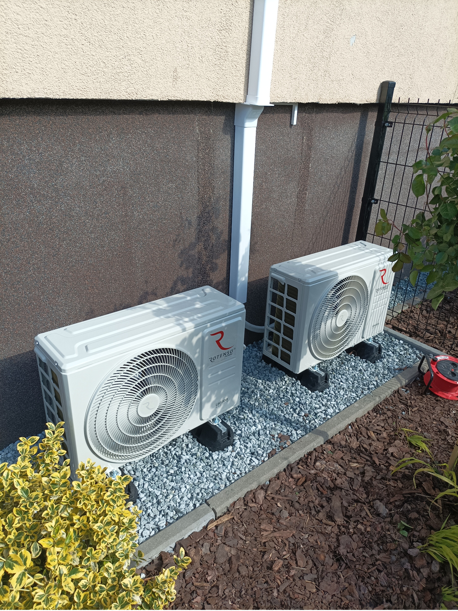 Dwa białe klimatyzatory Rotenso Ukura zamontowane na zewnątrz budynku, na podłożu z drobnych kamieni, obok rabaty z roślinami. Widoczny fragment elewacji.