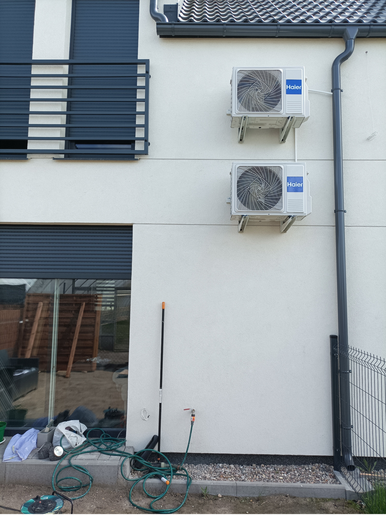 Dwa klimatyzatory Haier zamontowane na elewacji domu z szarymi okiennicami i rynną. W tle widoczny fragment ogrodu z wężem ogrodowym.