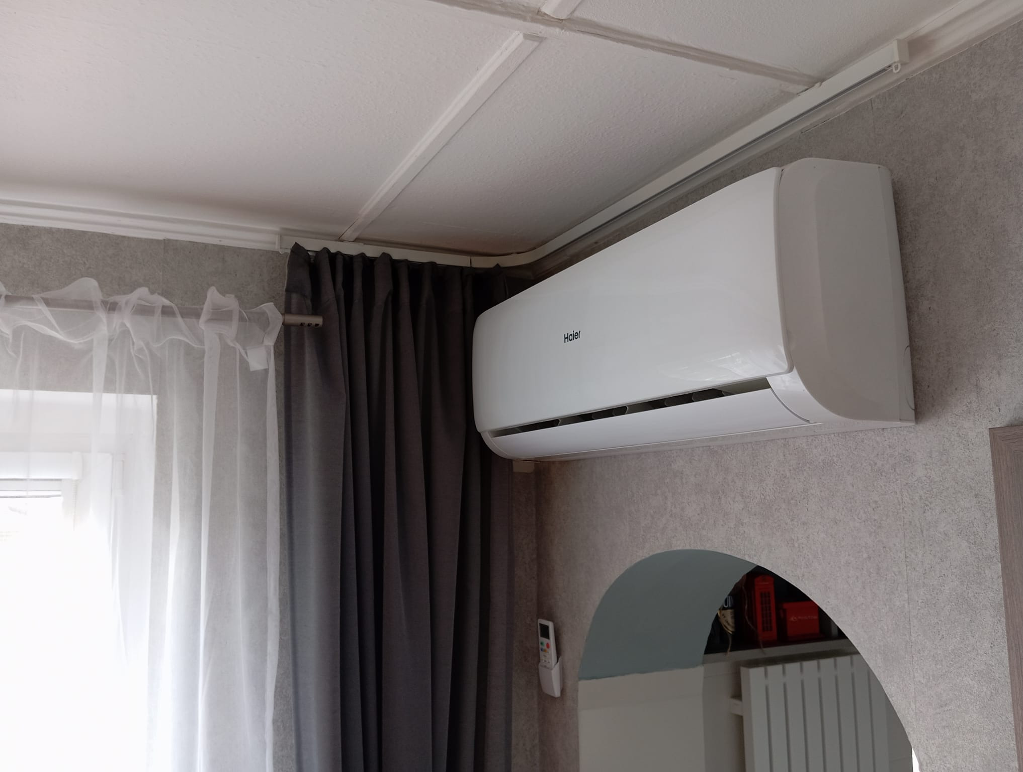 Biała klimatyzacja Haier na ścianie z szarą tapetą, obok okno z zasłonami. Widoczny pilot do klimatyzacji i fragment dekoracyjnego łuku w ścianie.
