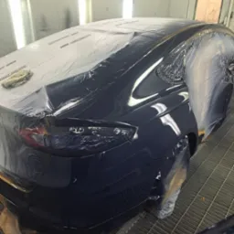 Błyszczący, granatowy samochód w lakierni, częściowo oklejony folią ochronną, przygotowany do polerowania. Widoczne odbicia światła na świeżo polakierowanej karoserii.