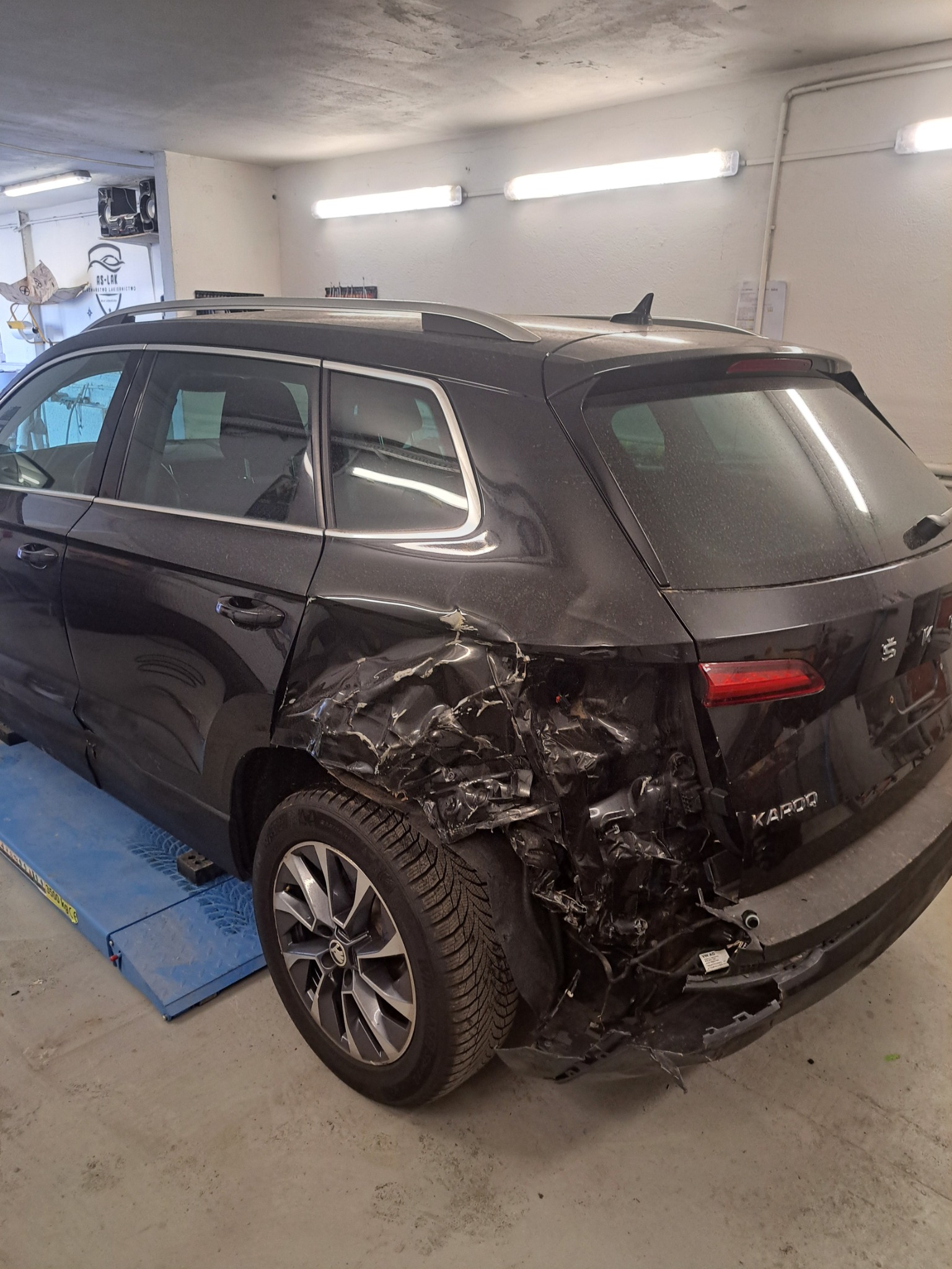Uszkodzony tył czarnej Skody Karoq na podnośniku warsztatowym. Widoczne zgniecione blachy i połamane elementy. W tle ściana warsztatu z oświetleniem.