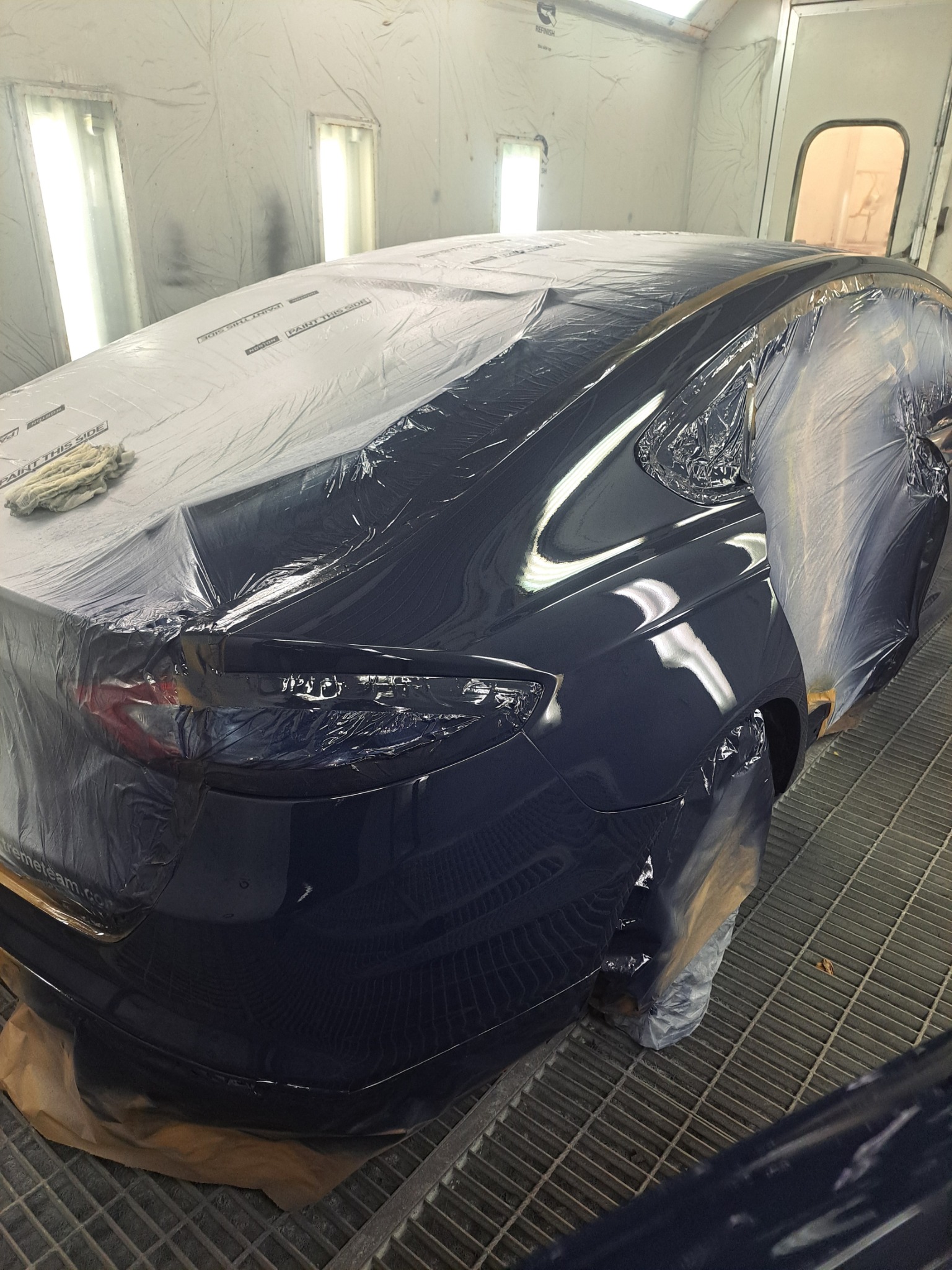 Błyszczący, granatowy samochód w lakierni, częściowo oklejony folią ochronną, przygotowany do polerowania. Widoczne odbicia światła na świeżo polakierowanej karoserii.