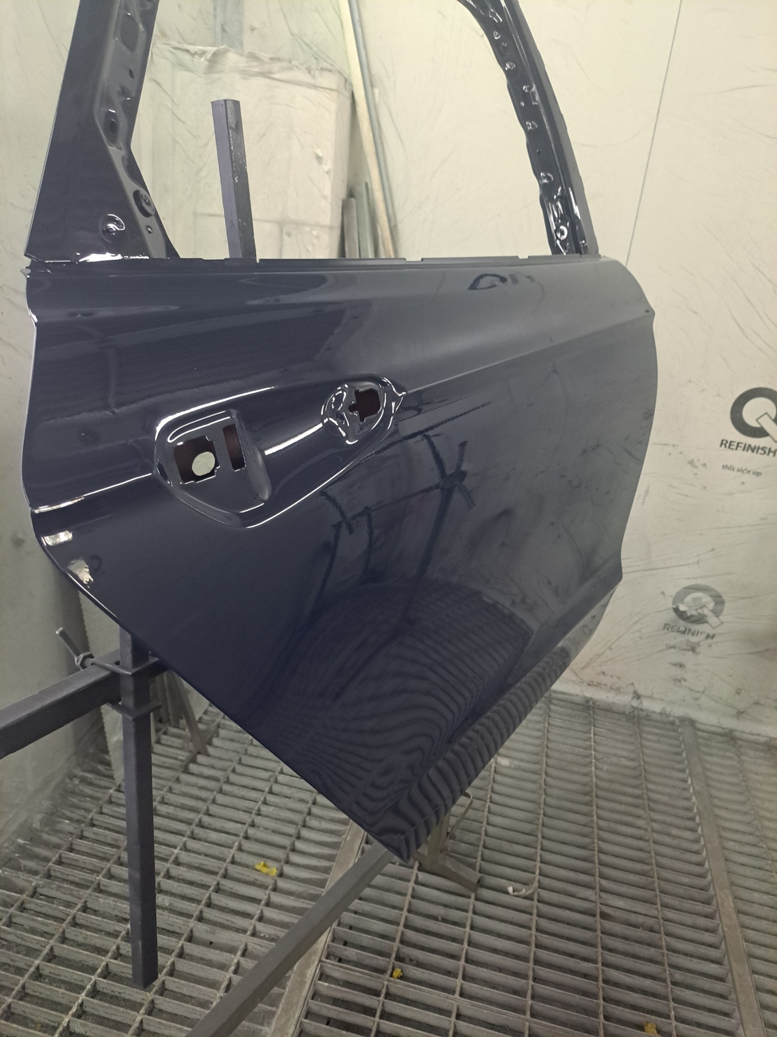 Błyszczące, granatowe drzwi samochodowe po lakierowaniu, zawieszone w lakierni na metalowym stojaku, z widoczną kratką podłogową i logo producenta farb.