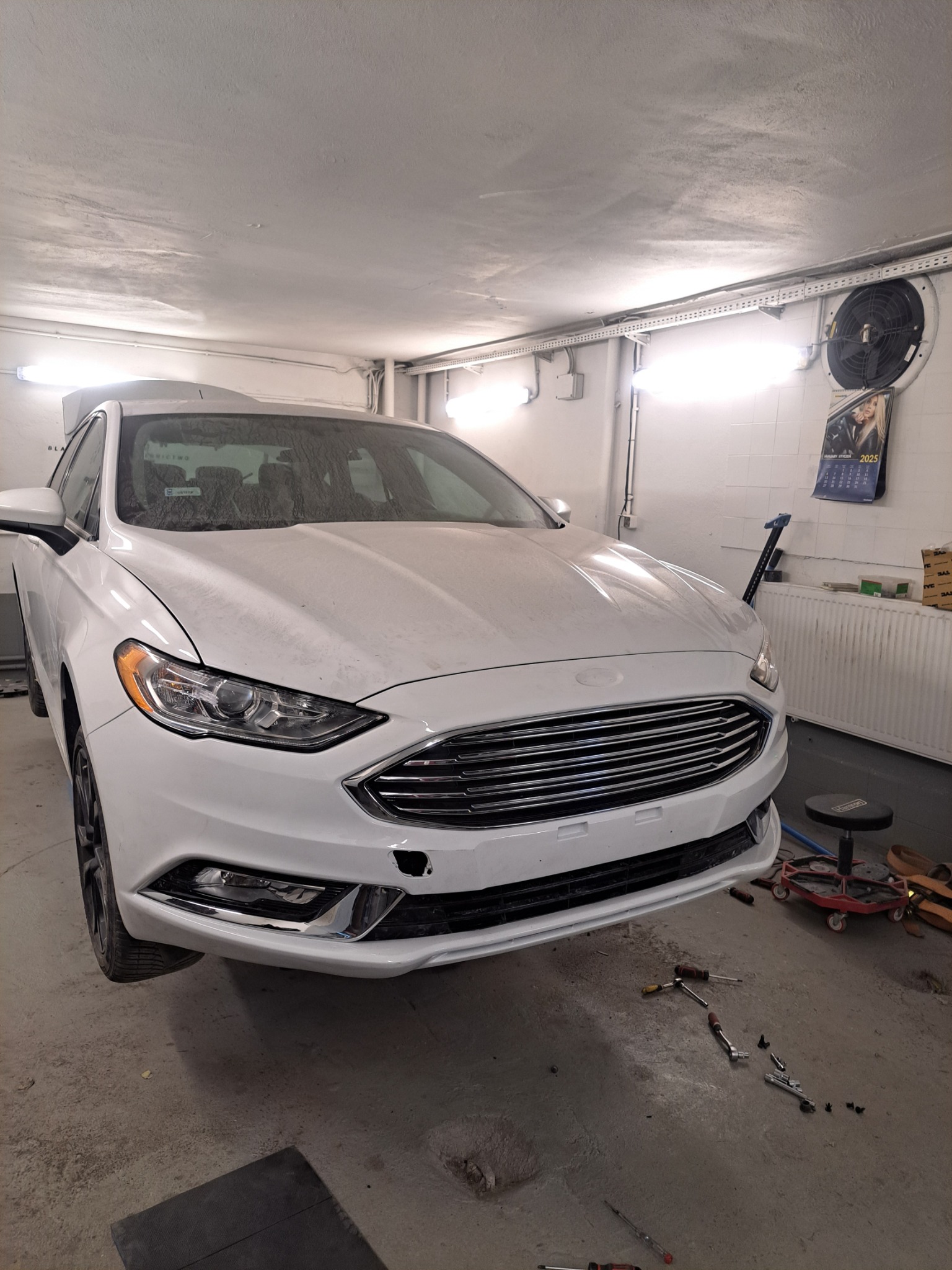 Biały Ford Fusion w warsztacie w Gdańsku przed naprawą. Widoczne narzędzia i uszkodzenia na przednim zderzaku. Auto uniesione, przygotowane do blacharsko-lakierniczych prac.