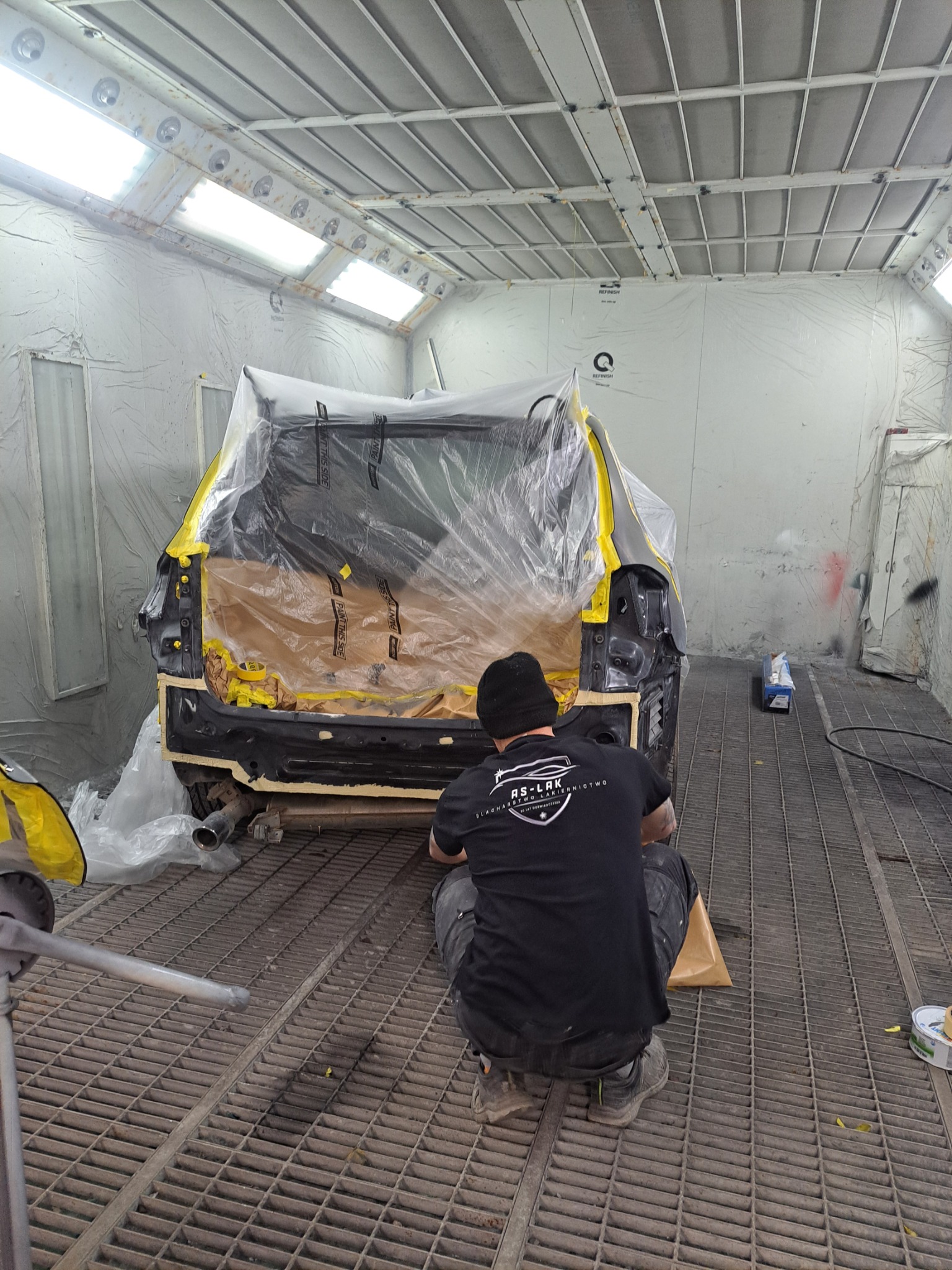 Przygotowanie auta do lakierowania w kabinie: pracownik w czarnej koszulce z logo firmy zabezpiecza karoserię folią i taśmą. Widoczny element wydechu i podłoga z kratą.