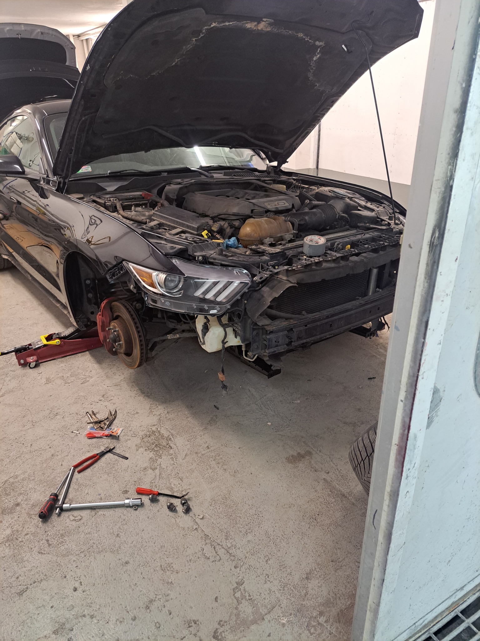 Czarny samochód z otwartą maską, bez przedniego zderzaka i jednego koła, stoi w warsztacie. Wokół leżą narzędzia.