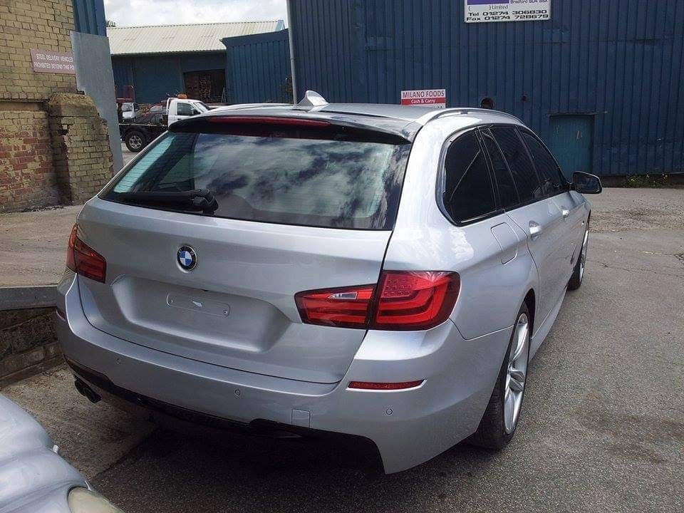 Srebrne BMW serii 5 kombi, widok z tyłu, zaparkowane na zewnątrz. Widoczne tylne światła LED i logo BMW. Tło stanowią budynki przemysłowe i częściowo widoczny inny pojazd.