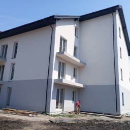 Lewon - Dwukolorowa elewacja budynku wielorodzinnego z balkonami, widok z dołu. Na pierwszym planie teren budowy i pracownik w czerwonej koszulce.