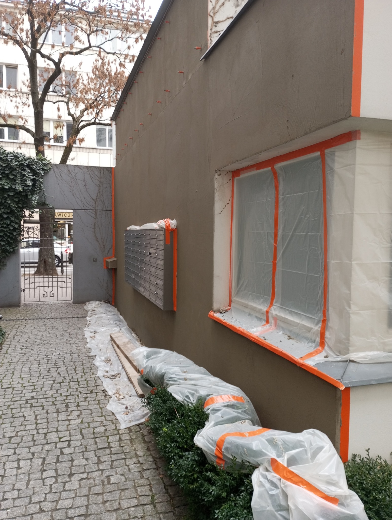 Elewacja budynku w trakcie tynkowania: świeży tynk, okna i elementy zabezpieczone folią i taśmą. Widoczne skrzynki pocztowe i fragment chodnika z kostki.