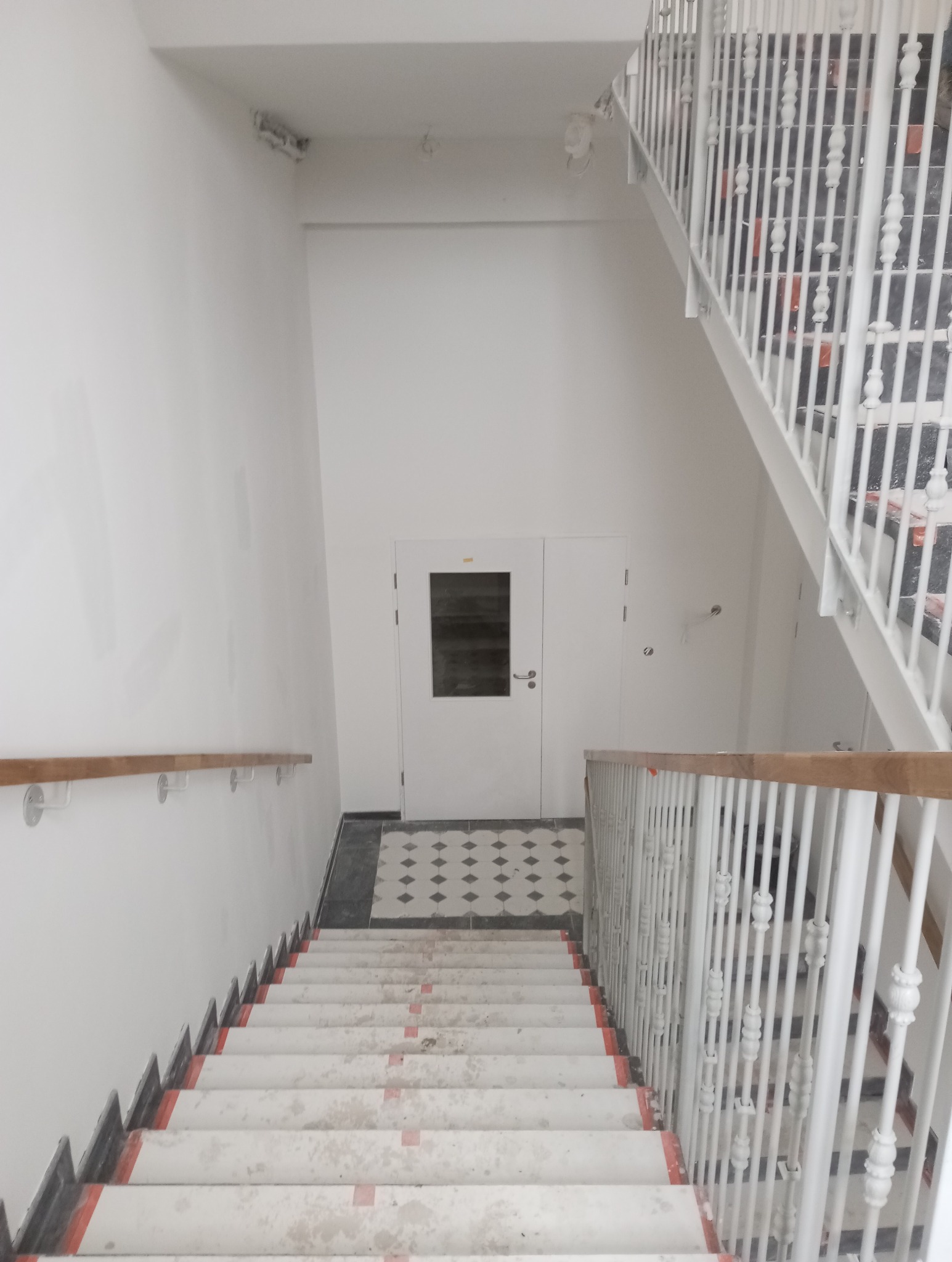 Białe schody z drewnianą poręczą i metalowymi barierkami w trakcie wykończenia. Widoczne zabezpieczenia na schodach i podłodze. Ściany pomalowane na biało.