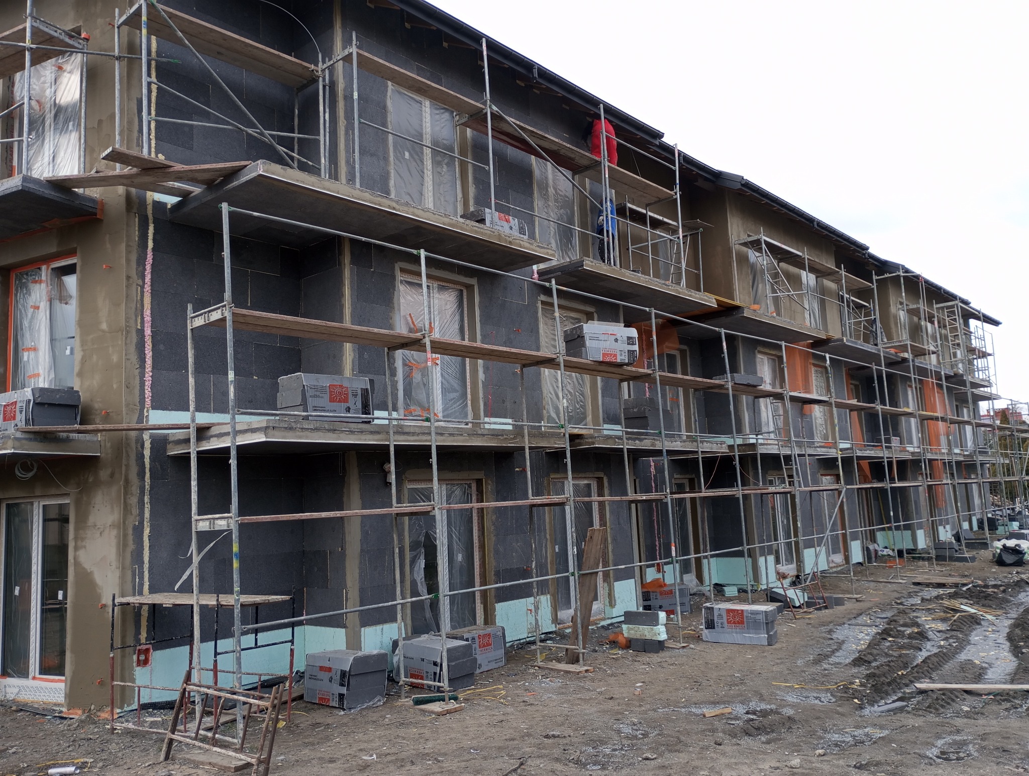 Budynek w trakcie ocieplania, widoczne rusztowania i materiały izolacyjne. Częściowo otynkowana elewacja. Na rusztowaniach leżą paczki z materiałem izolacyjnym.