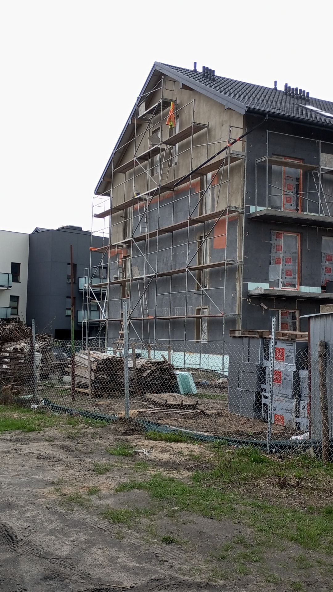 Budynek wielorodzinny w trakcie ocieplania, widoczne rusztowania i materiały izolacyjne. Częściowo otynkowana elewacja, okna zabezpieczone folią.