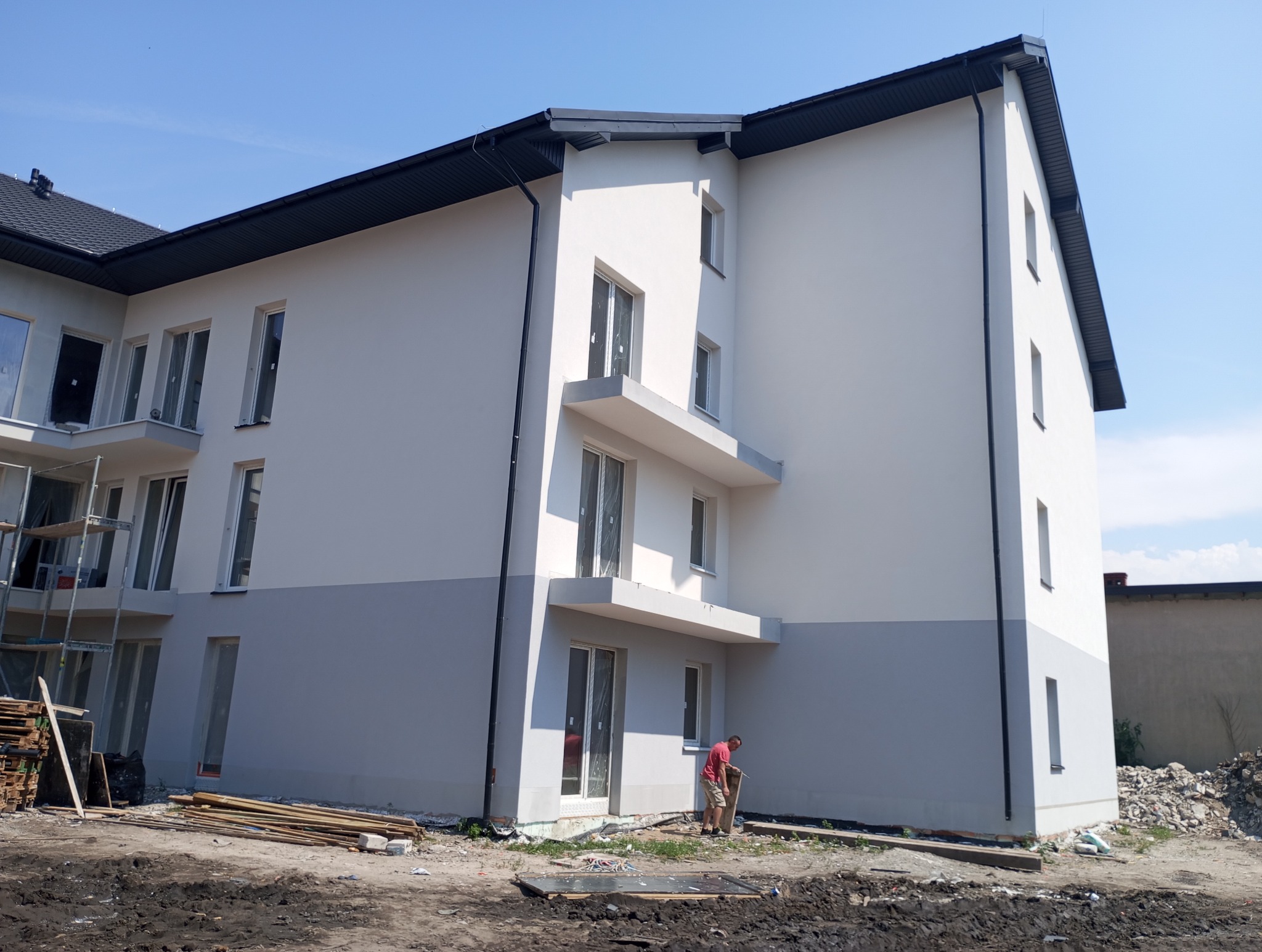 Dwukolorowa elewacja budynku wielorodzinnego z balkonami, widok z dołu. Na pierwszym planie teren budowy i pracownik w czerwonej koszulce.