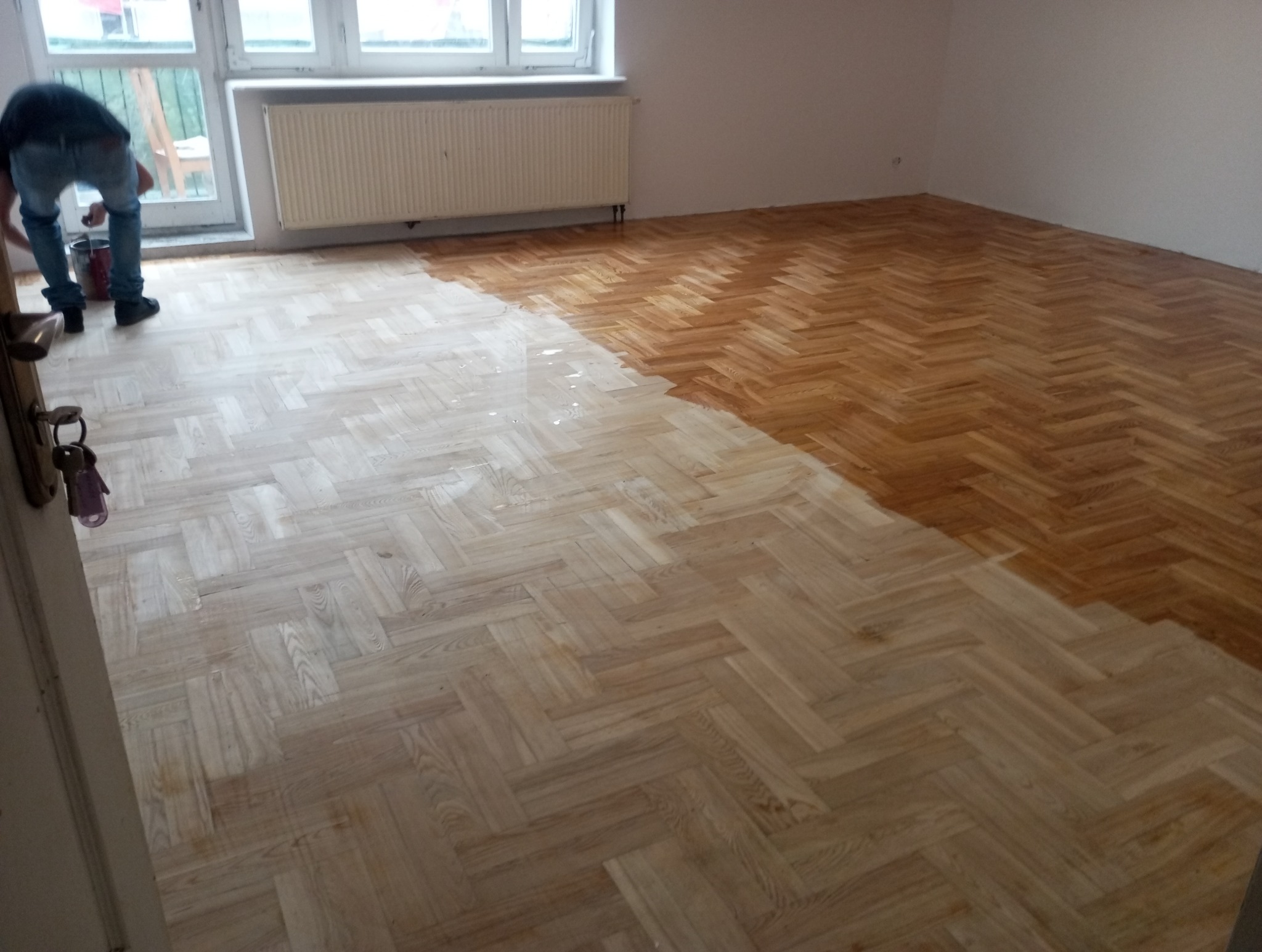 Parkiet w jodełkę częściowo polakierowany. Widoczny fragment osoby lakierującej podłogę i wiadro z lakierem. Okno i grzejnik w tle. Podłoga w trakcie renowacji.