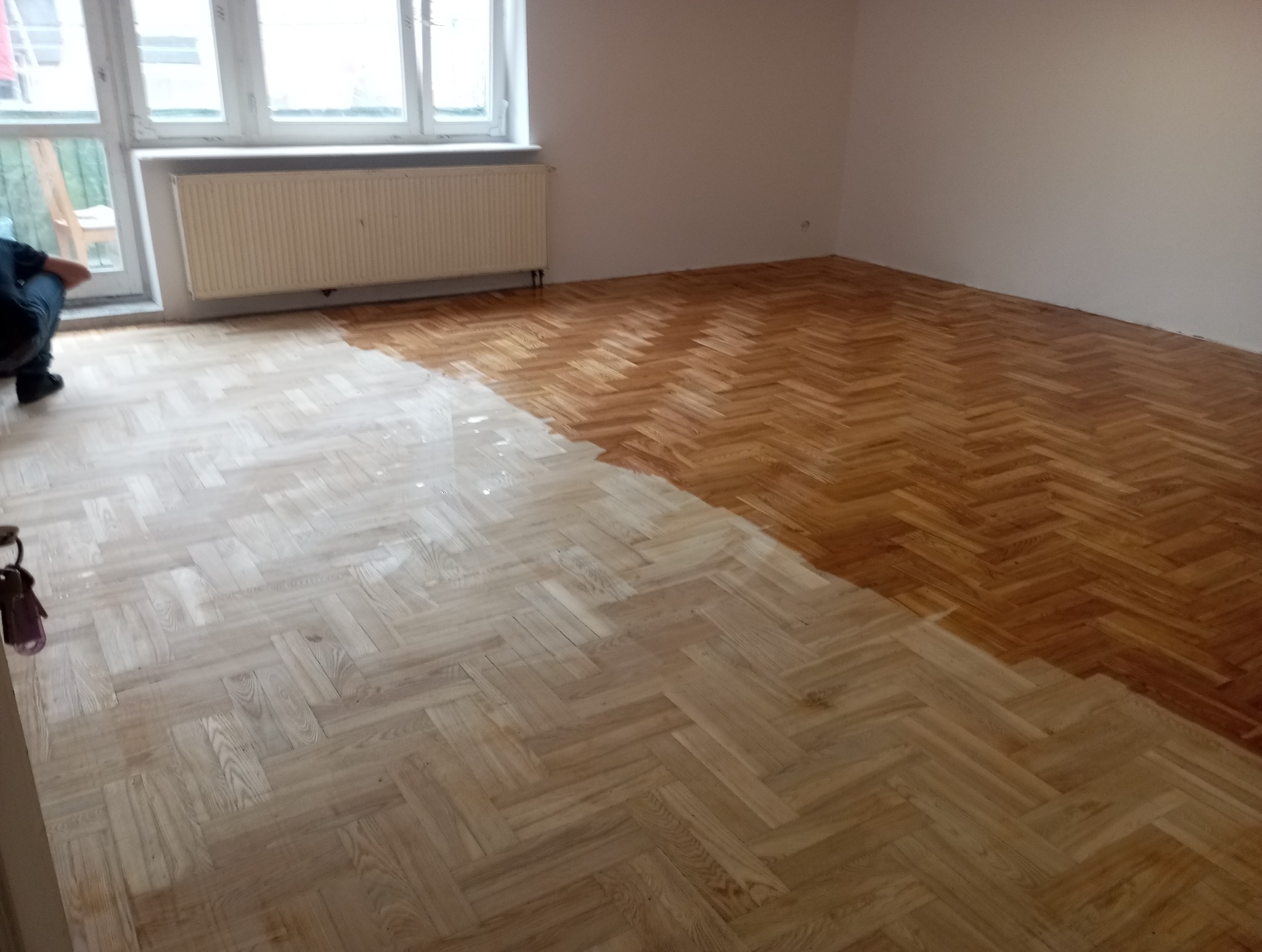 Parkiet w jodełkę po renowacji: połowa powierzchni wycyklinowana i polakierowana, druga połowa surowa. Widoczny fragment osoby. Okno z grzejnikiem w tle.