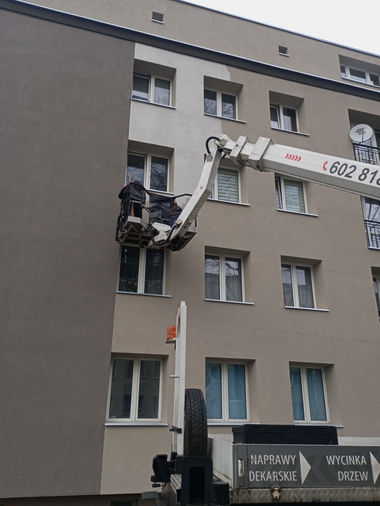 Prace elewacyjne na wysokościach: platforma podnośnika przy budynku, fragment pomalowanej ściany, widoczne okna. Naprawy dekarskie i wycinka drzew na pojeździe.