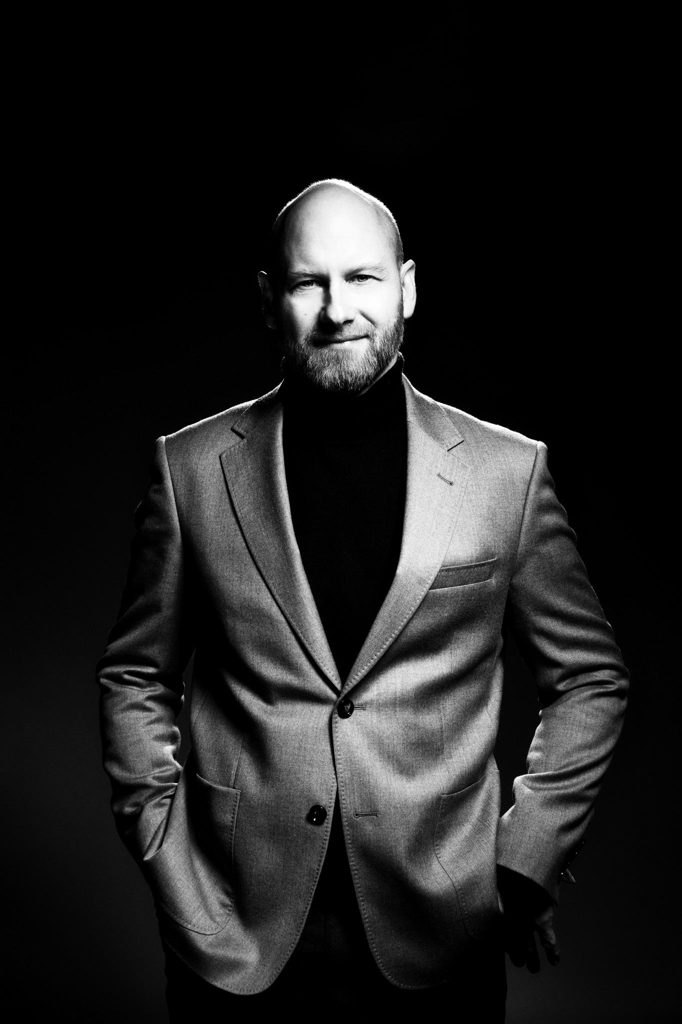 Czarno-biały portret mężczyzny z brodą, ubranego w golf i marynarkę, z rękami w kieszeniach, na ciemnym tle, z mocnym oświetleniem bocznym.