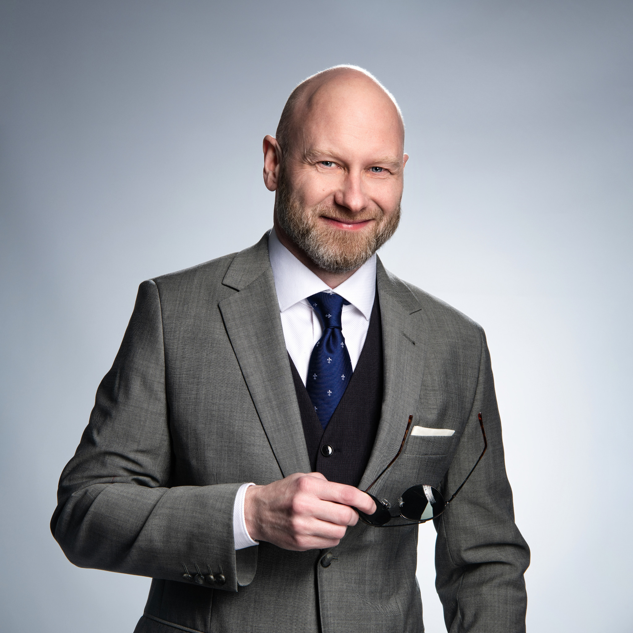 Portret uśmiechniętego mężczyzny w szarym garniturze, z brodą i okularami w dłoni, na jasnym tle. Profesjonalny wizerunek.