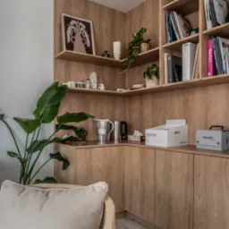 Kącik biurowy z drewnianymi półkami i szafkami, dekoracjami, roślinami i krzesłem z poduszką. Ciepłe oświetlenie podkreśla naturalny odcień drewna.