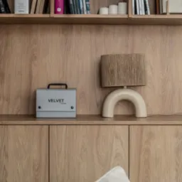 Drewniana półka z książkami, lampą w stylu retro i szarą skrzynką Velvet I-orner. Minimalistyczny design wnętrza, ciepłe oświetlenie.