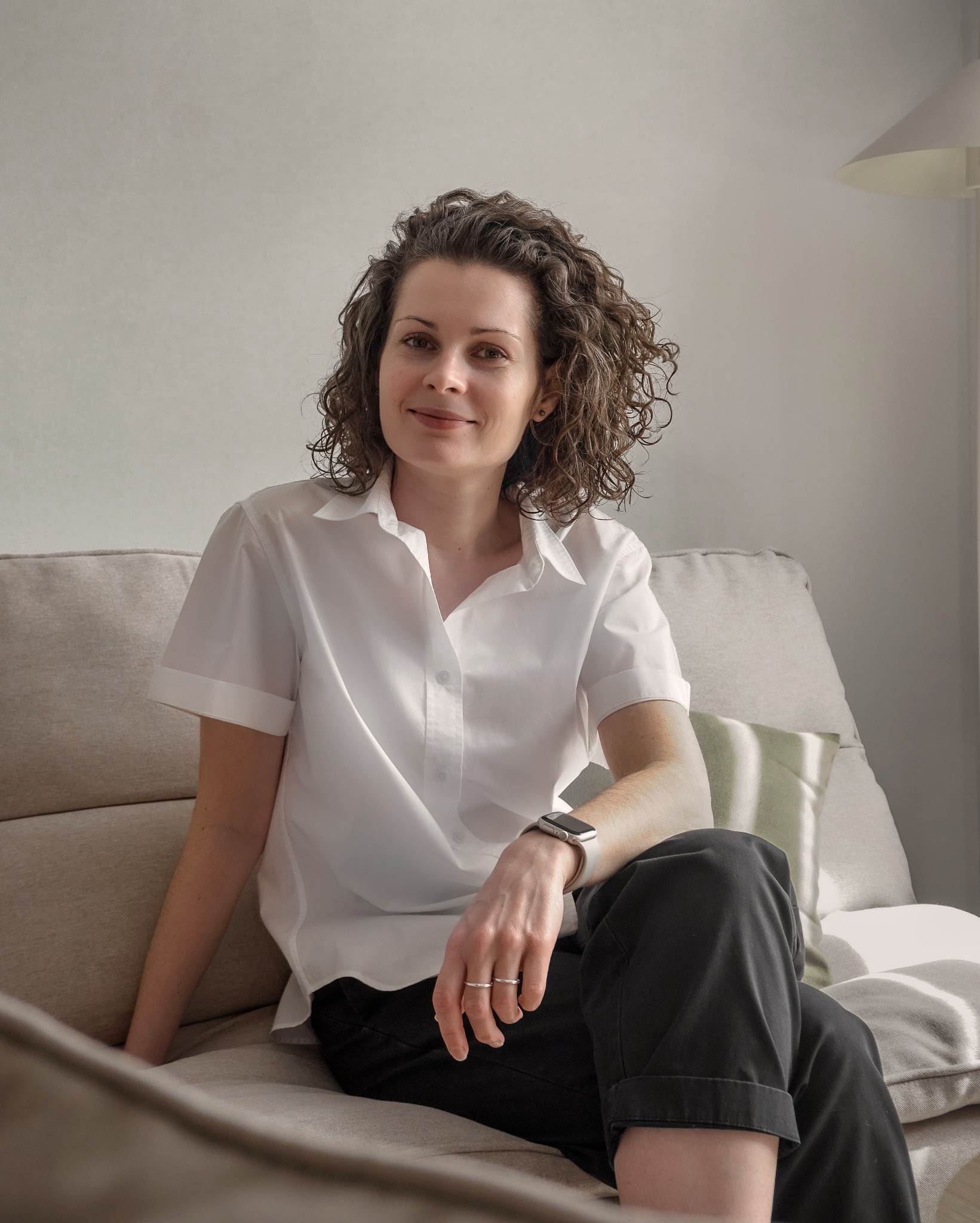 Portret kobiety z kręconymi włosami siedzącej na beżowej sofie w białej koszuli i ciemnych spodniach. W tle widoczna lampa i jasna ściana.
