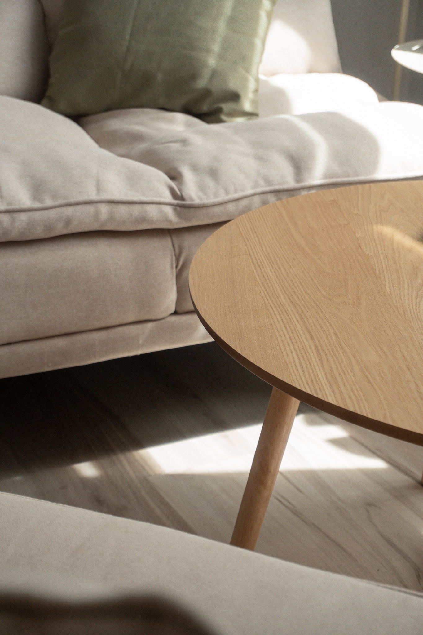 Jasny salon z beżową sofą, zieloną poduszką i okrągłym, drewnianym stolikiem kawowym w minimalistycznym stylu, oświetlonym naturalnym światłem.