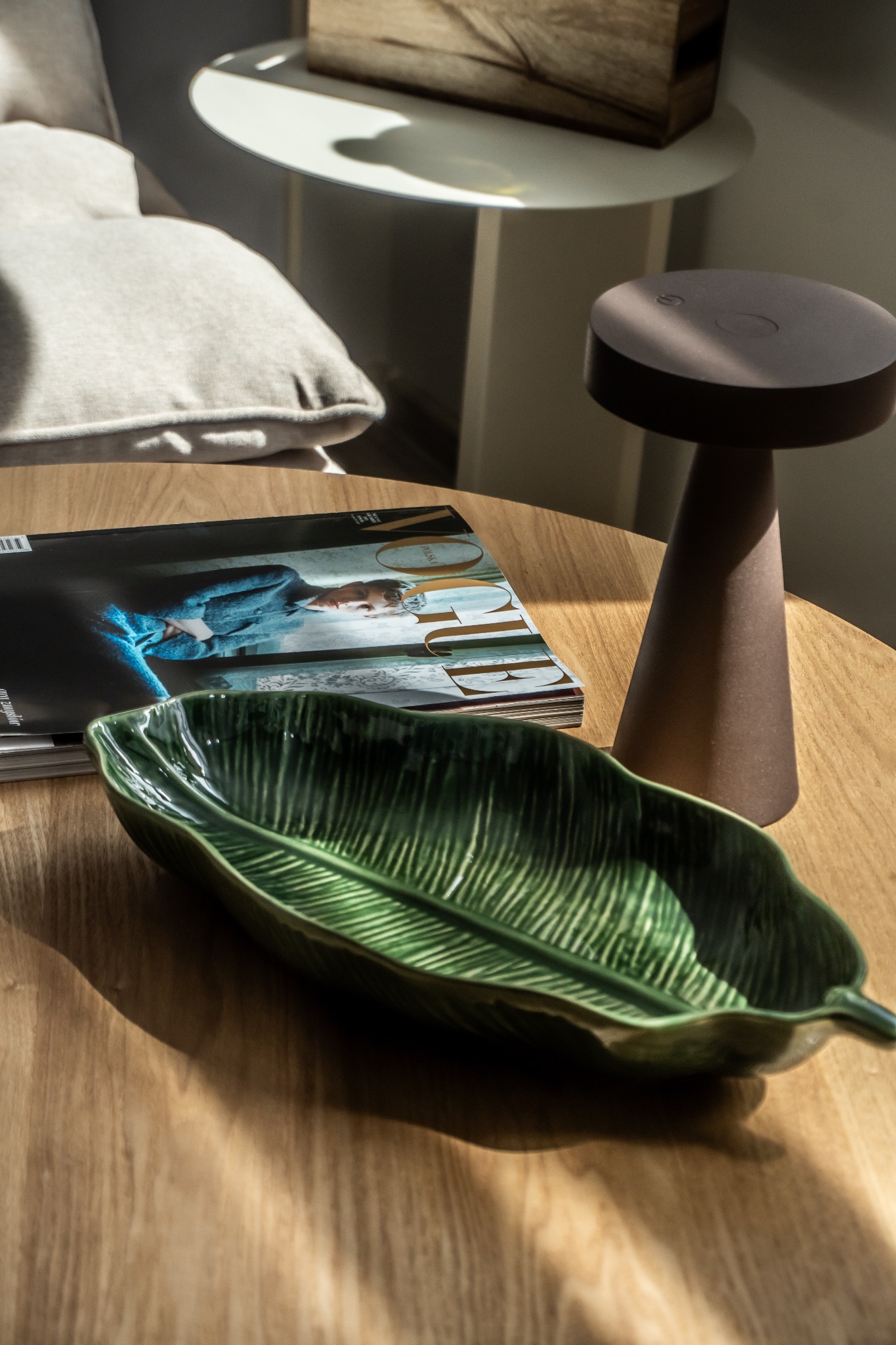 Dekoracyjny stół z ceramiczną paterą w kształcie liścia, magazynem modowym i nowoczesną lampką. Elementy aranżacji wnętrza w ciepłym oświetleniu.