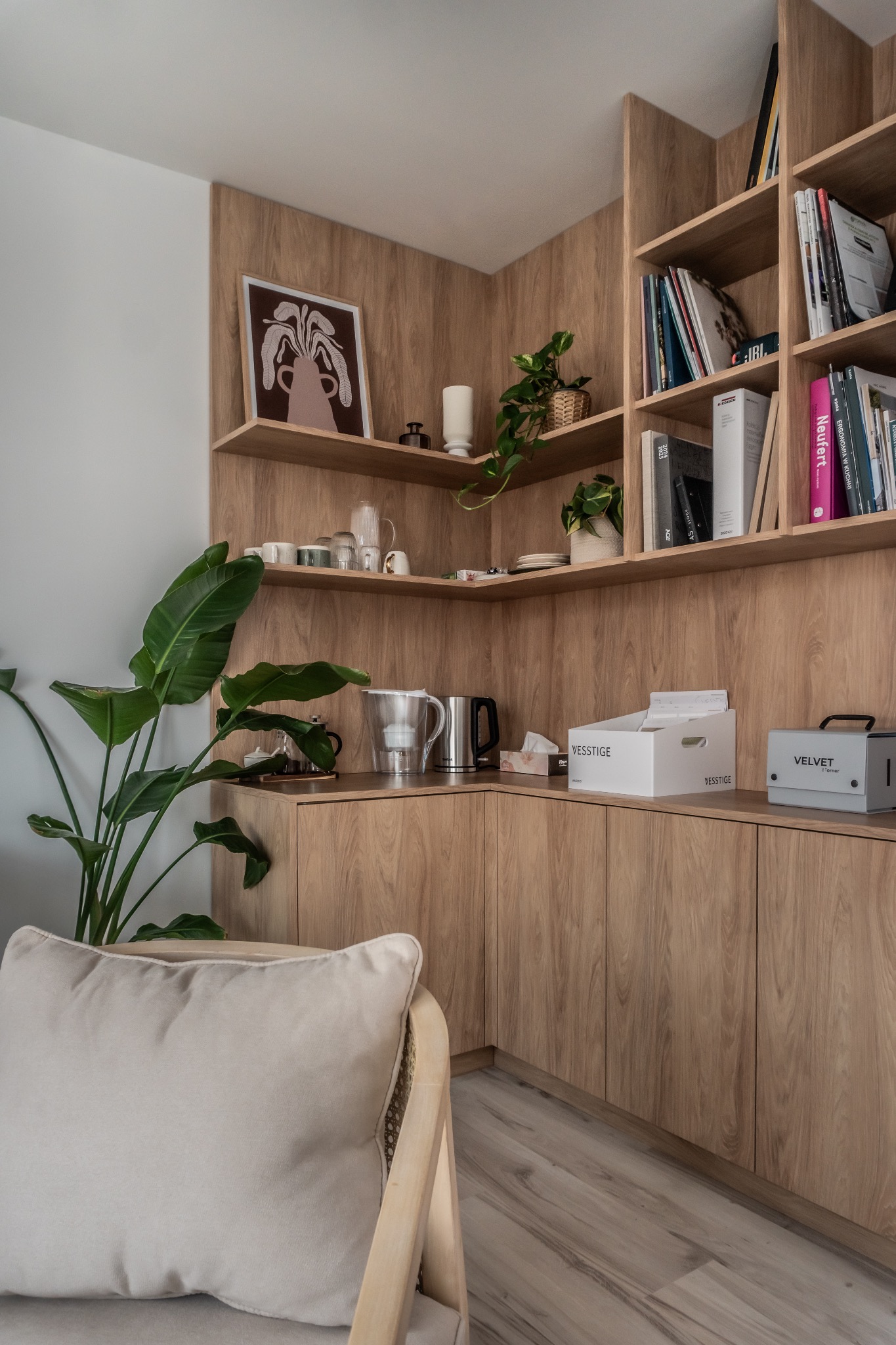 Kącik biurowy z drewnianymi półkami i szafkami, dekoracjami, roślinami i krzesłem z poduszką. Ciepłe oświetlenie podkreśla naturalny odcień drewna.
