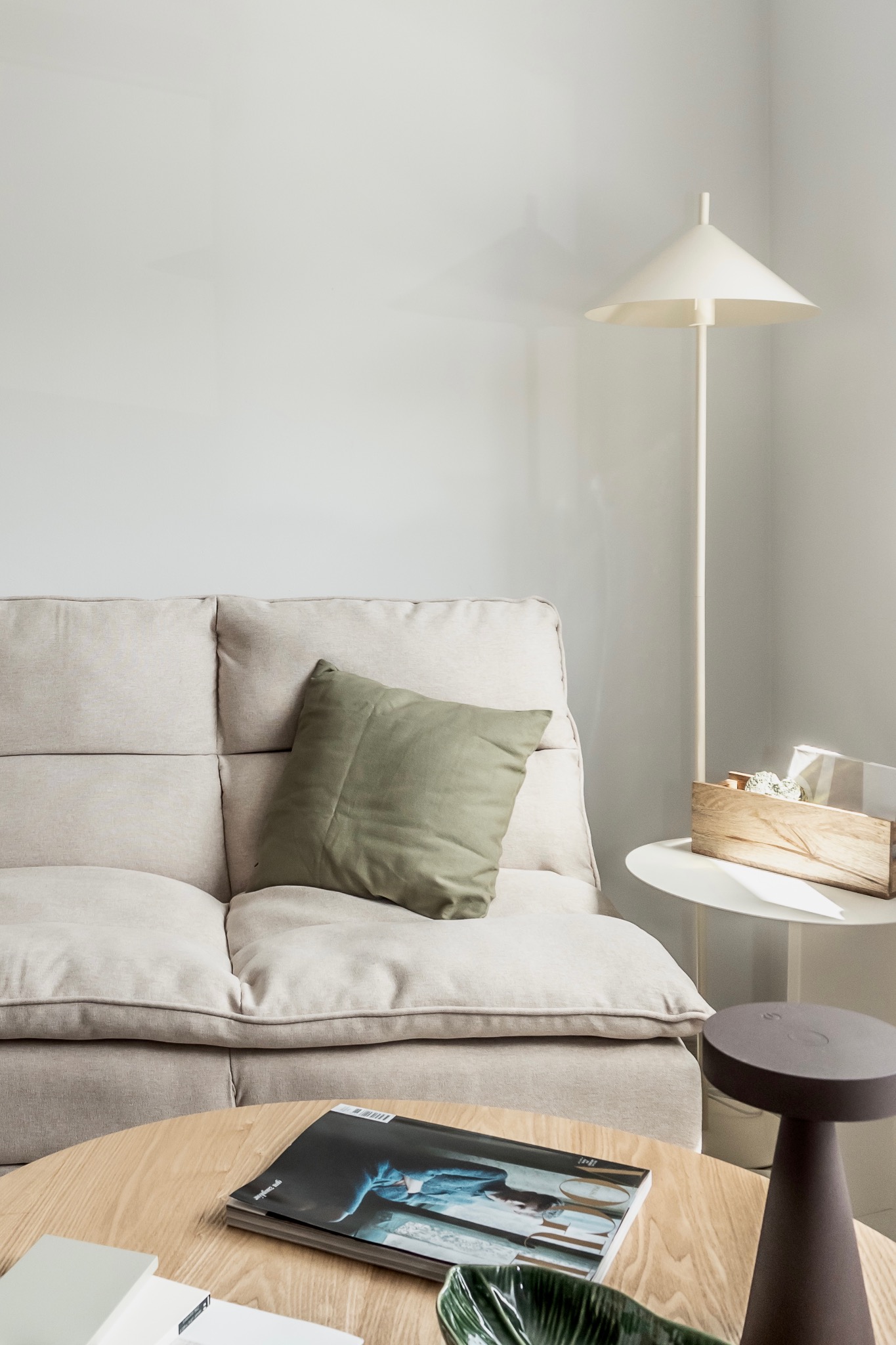 Jasny salon z beżową sofą, zieloną poduszką, drewnianym stolikiem kawowym i lampą podłogową. Na stoliku magazyn i dekoracje. Minimalistyczny wystrój.