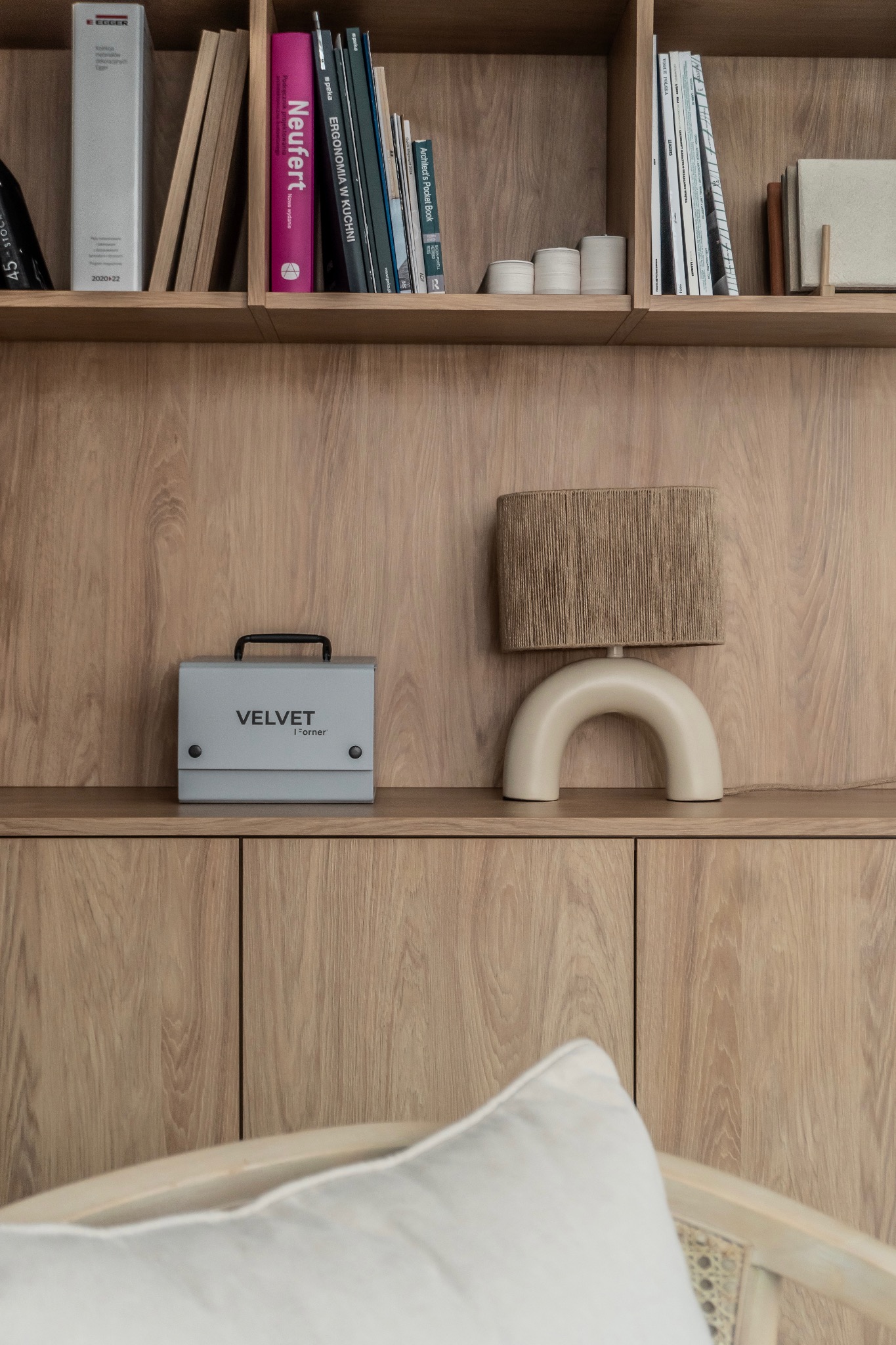 Drewniana półka z książkami, lampą w stylu retro i szarą skrzynką Velvet I-orner. Minimalistyczny design wnętrza, ciepłe oświetlenie.
