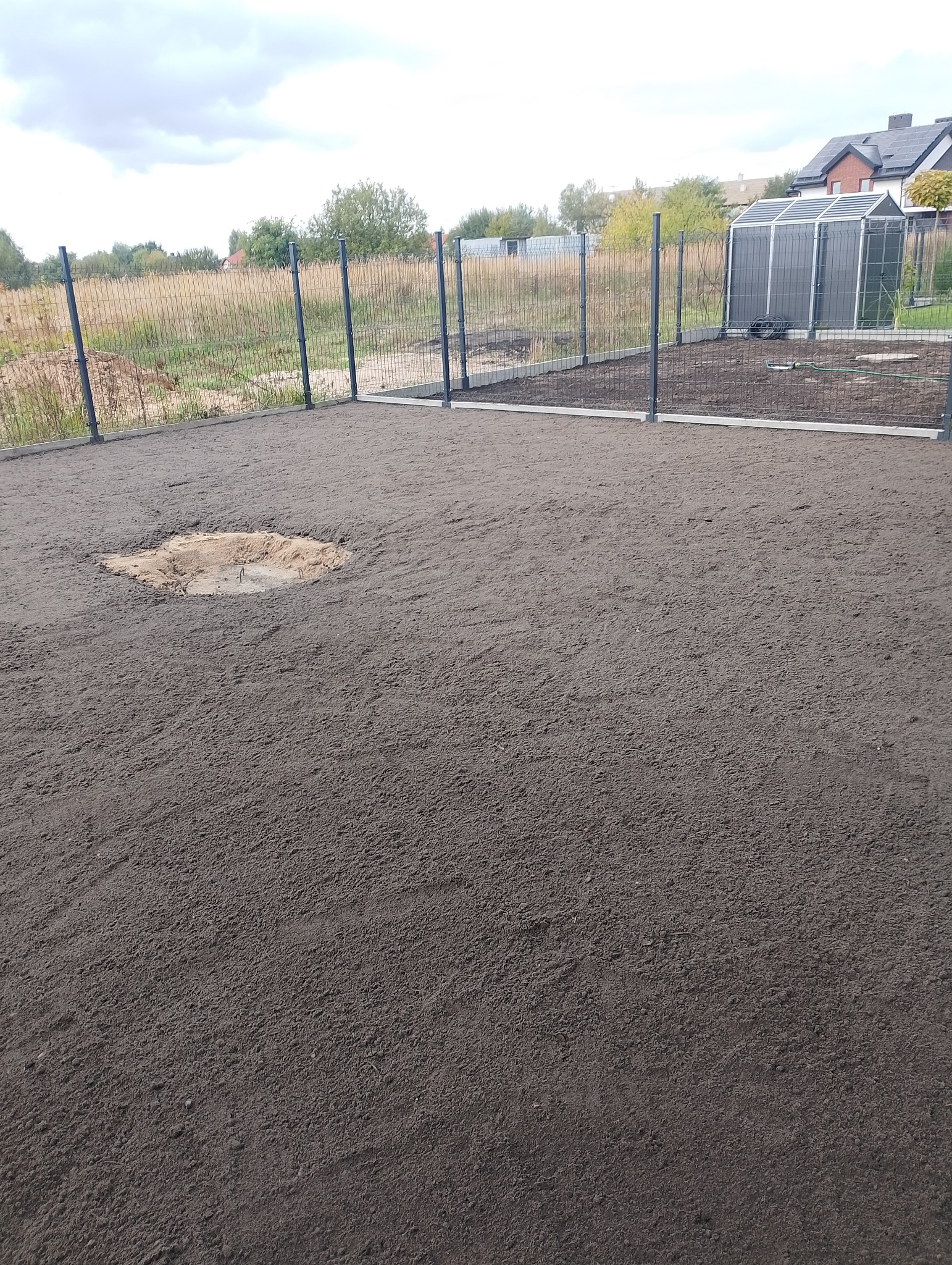 Przygotowany teren pod trawnik, wyrównana ziemia z widocznymi śladami po bronowaniu. W tle ogrodzenie panelowe i szklarnia.