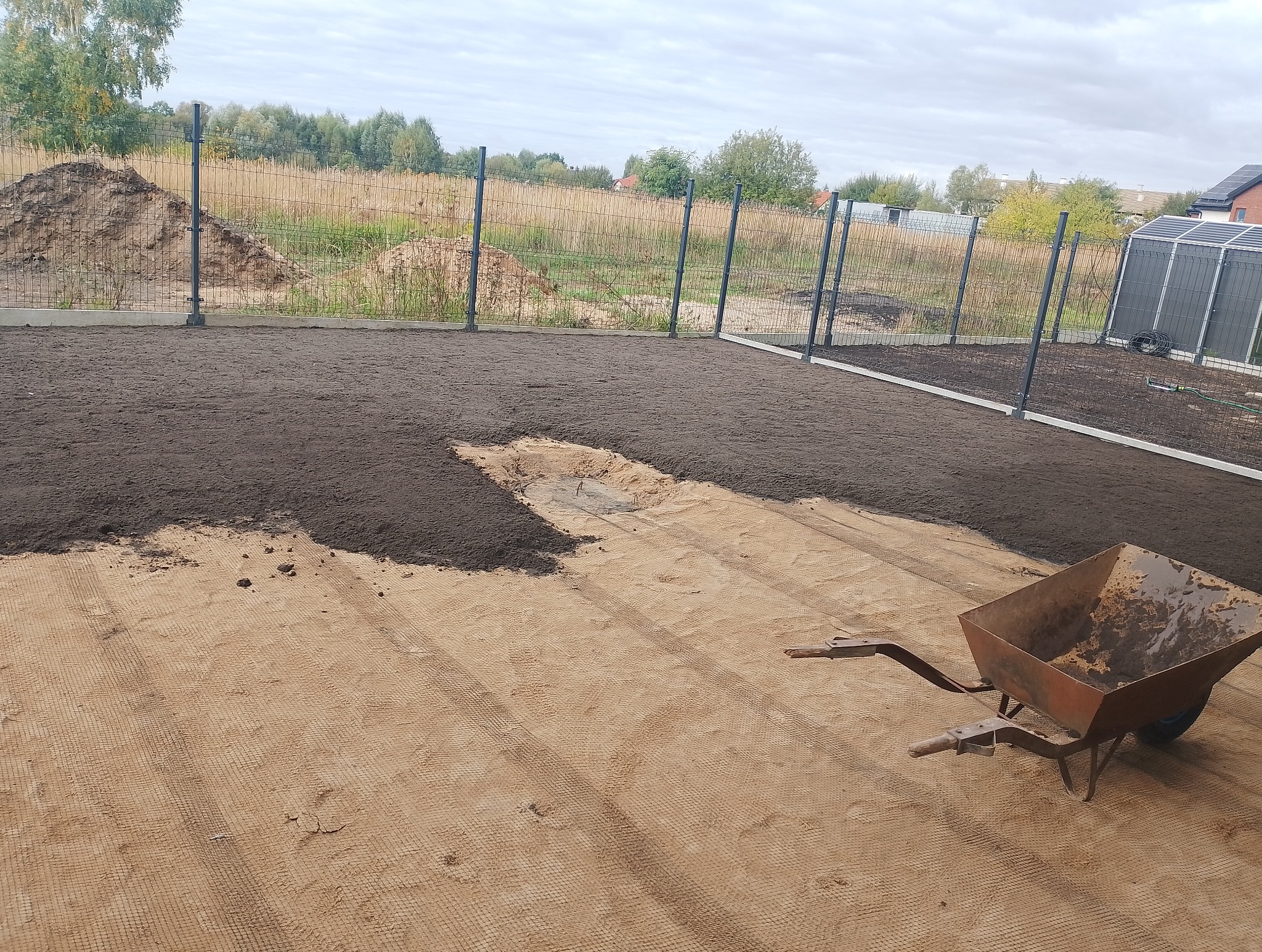 Przygotowanie terenu pod trawnik: rozplantowana ziemia, siatka wzmacniająca i taczka. Widoczne ogrodzenie panelowe i pryzma ziemi w tle.