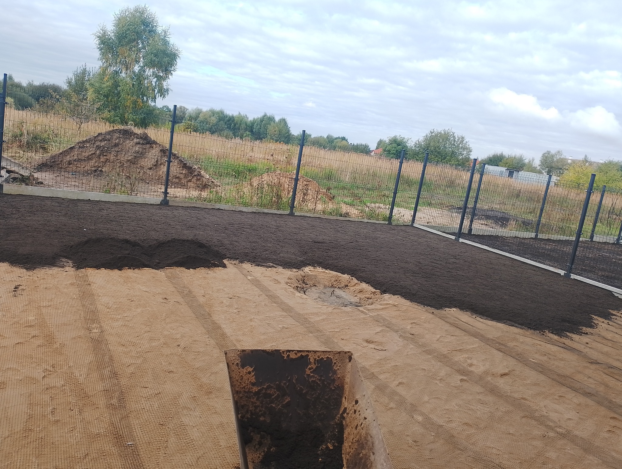 Przygotowanie terenu pod trawnik: wyrównywanie ziemi i rozsypywanie ciemnej gleby na podłożu z siatką, widoczny fragment ogrodzenia i kopiec ziemi w tle.