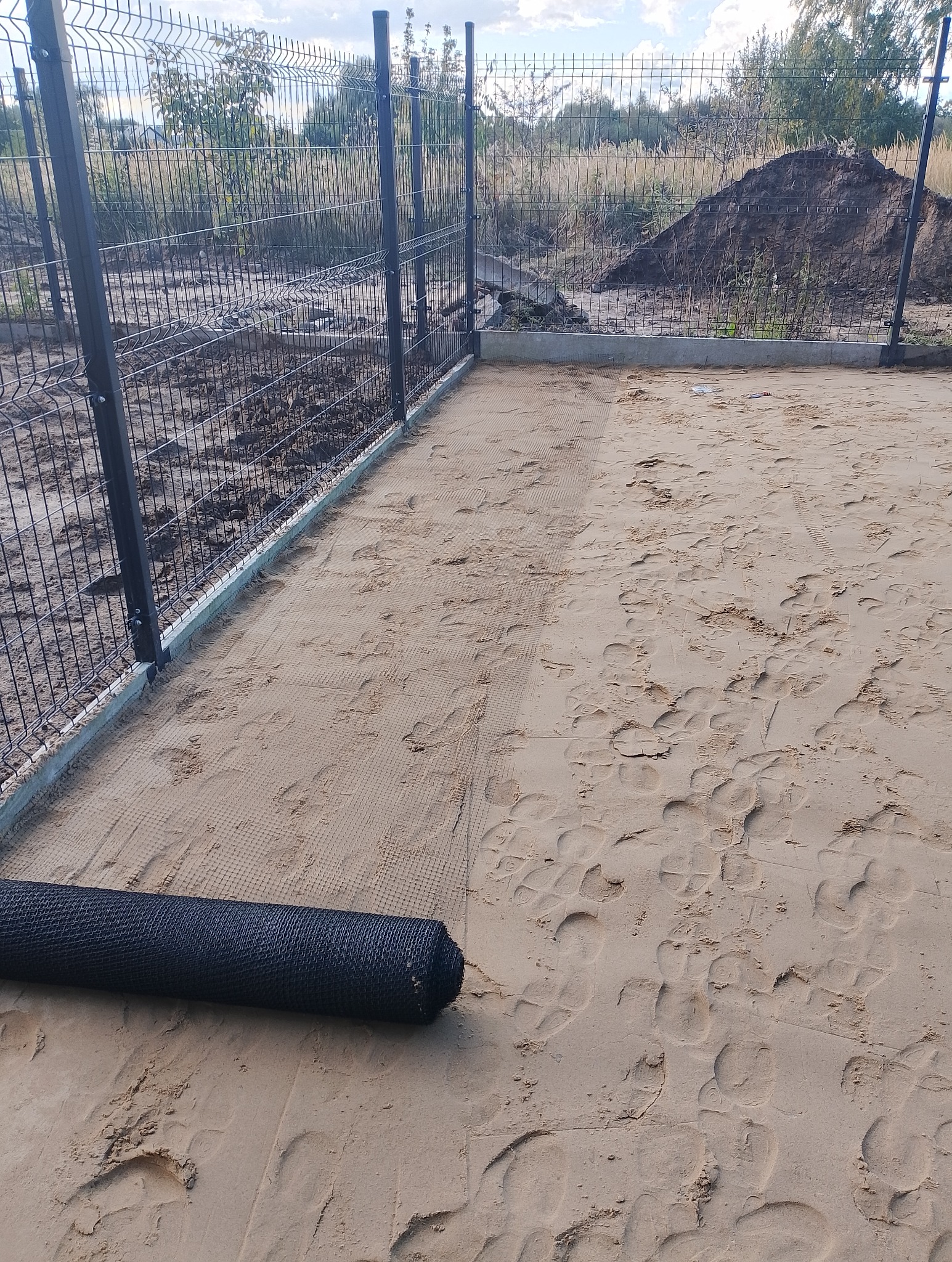 Przygotowany teren pod trawnik z rolki, widoczna siatka wzmacniająca i zwinięta rolka trawy syntetycznej. W tle metalowe ogrodzenie i pryzma ziemi.