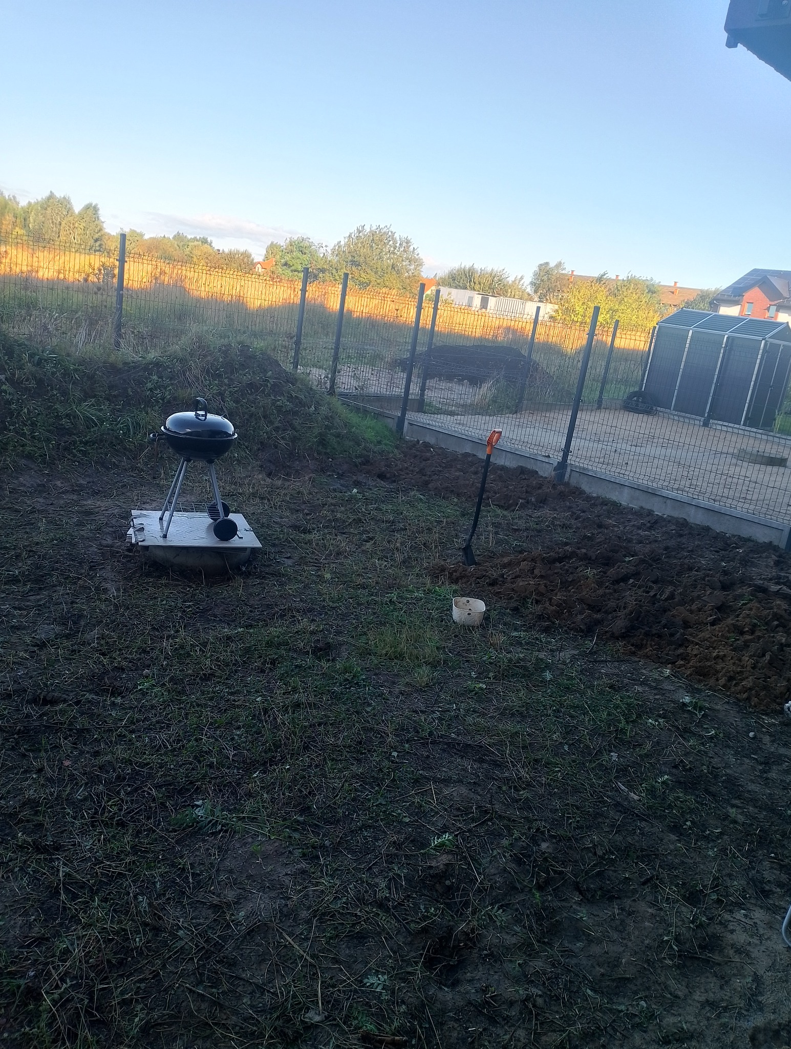 Przygotowanie terenu pod trawnik: rozkopana ziemia, szpadel wbity w ziemię. W tle ogrodzenie i grill na betonowej płycie. Poranne światło.