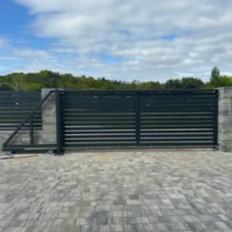 Nowoczesna, przesuwna brama wjazdowa z poziomymi panelami, szara kostka brukowa i słupki z imitacji kamienia. Widok z zewnątrz na tle zieleni.
