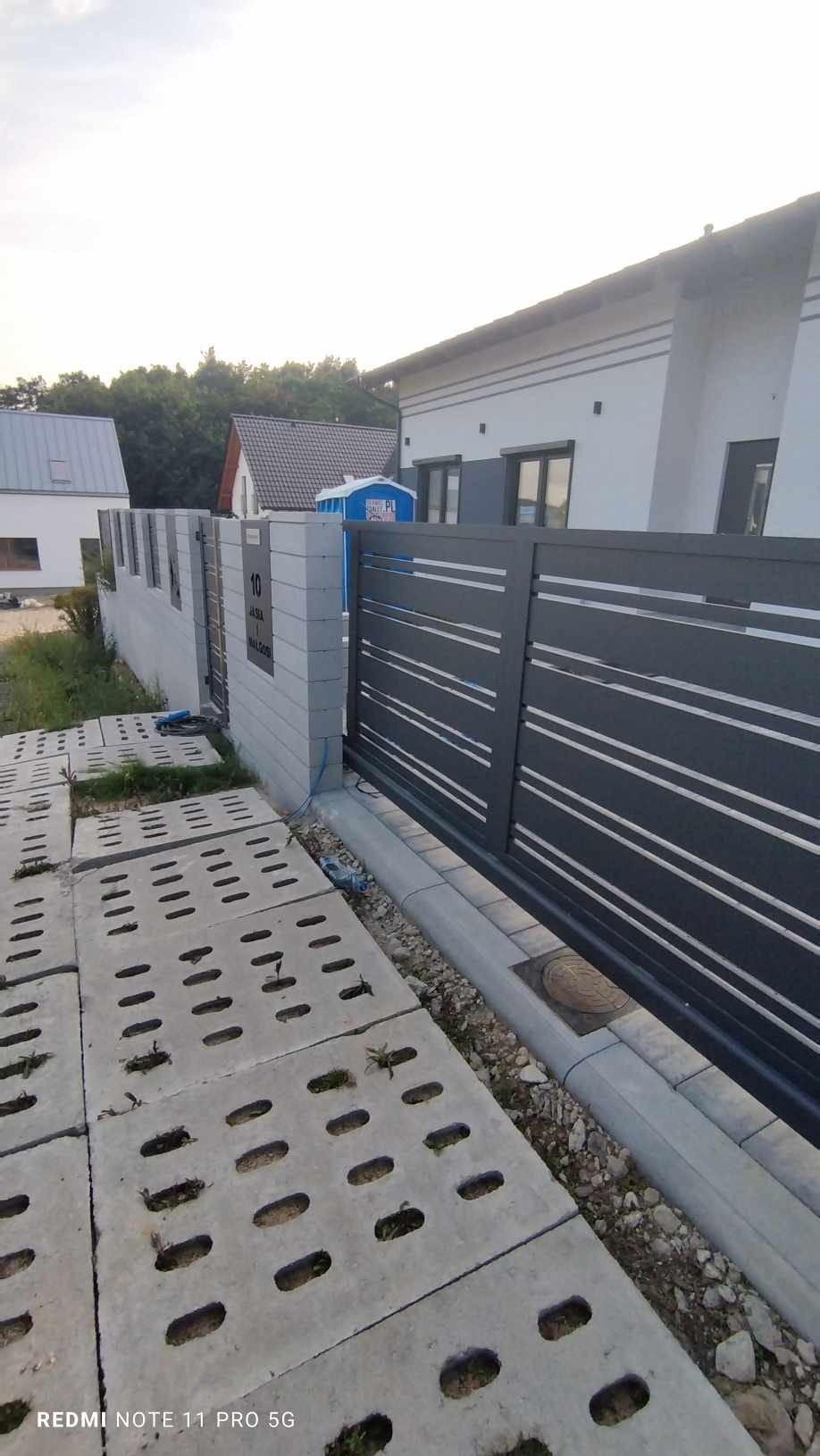 Nowoczesna, przesuwna brama wjazdowa z metalowymi panelami i betonowym ogrodzeniem z numerem domu, widok z zewnątrz na podjazd wyłożony ażurowymi płytami.