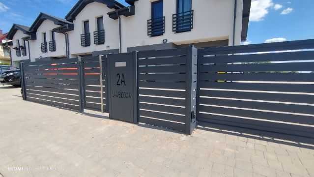 Nowoczesna, pozioma brama wjazdowa i furtka w kolorze grafitowym, z numerem posesji '2A' i nazwą ulicy, na tle jasnej elewacji domu.