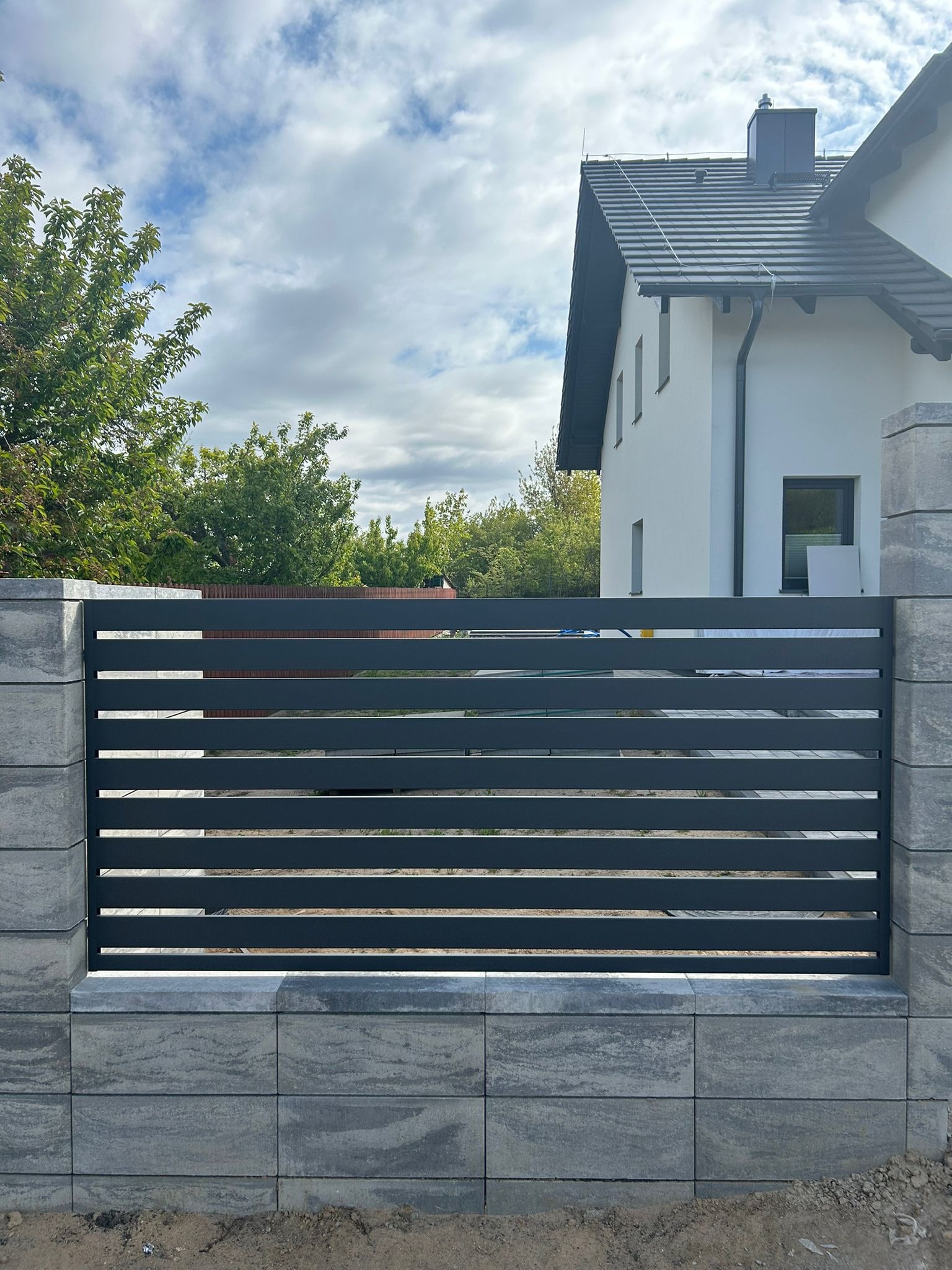 Nowoczesne ogrodzenie panelowe z poziomych sztachet w kolorze grafitowym, osadzone na słupkach z szarej kostki. W tle widoczny fragment domu jednorodzinnego.