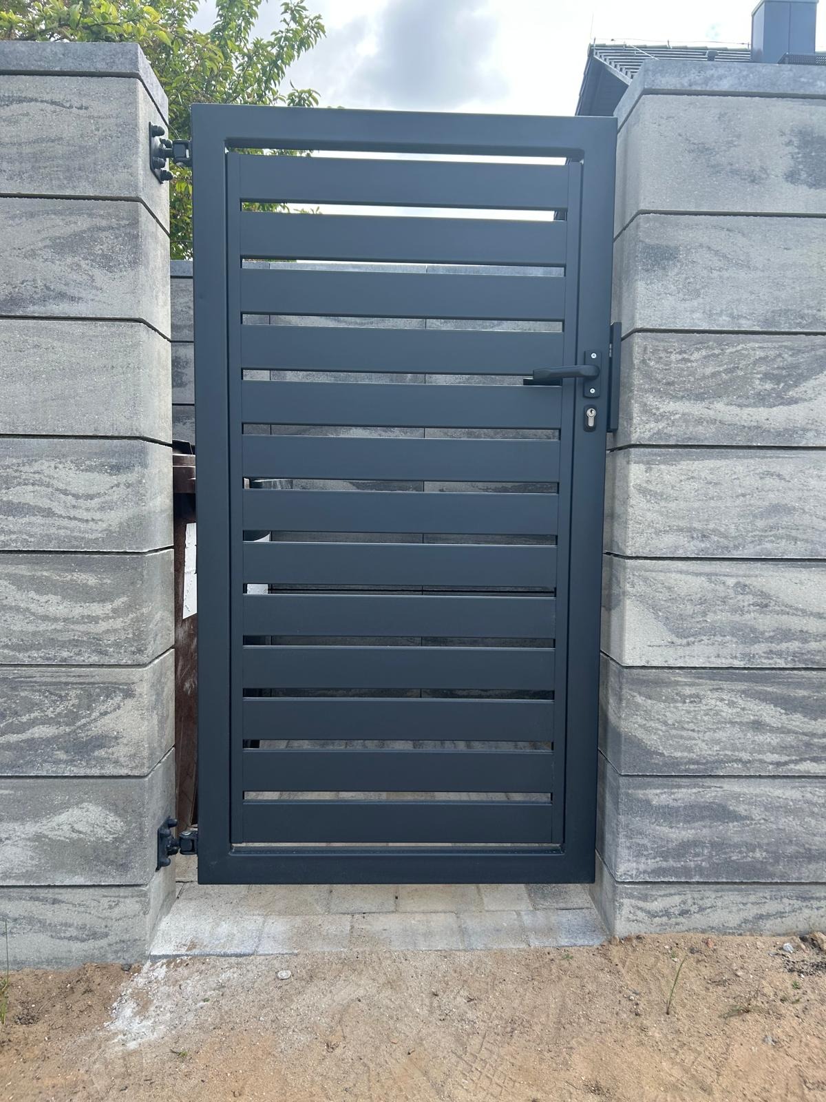 Nowoczesna, antracytowa furtka z poziomych profili, zamontowana na słupkach z szarej kostki betonowej o fakturze kamienia. Widoczne zawiasy i klamka.