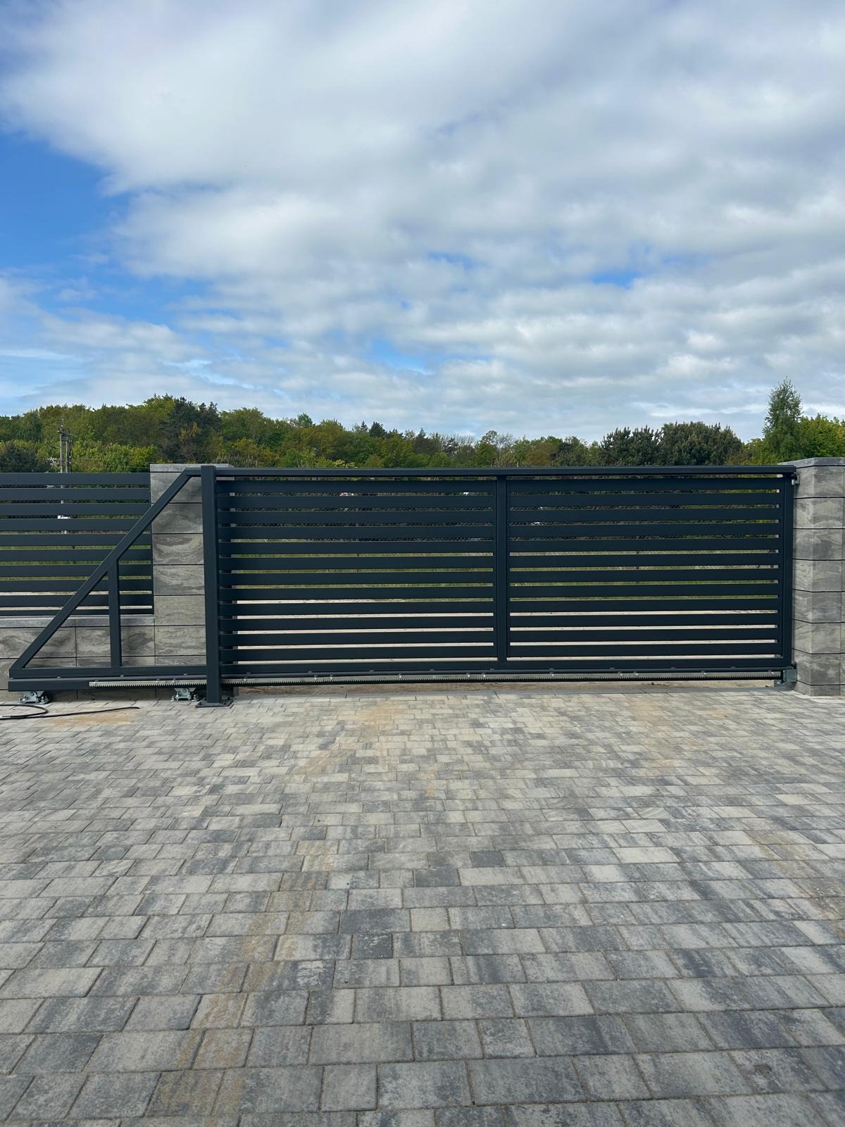 Nowoczesna, przesuwna brama wjazdowa z poziomymi panelami, szara kostka brukowa i słupki z imitacji kamienia. Widok z zewnątrz na tle zieleni.