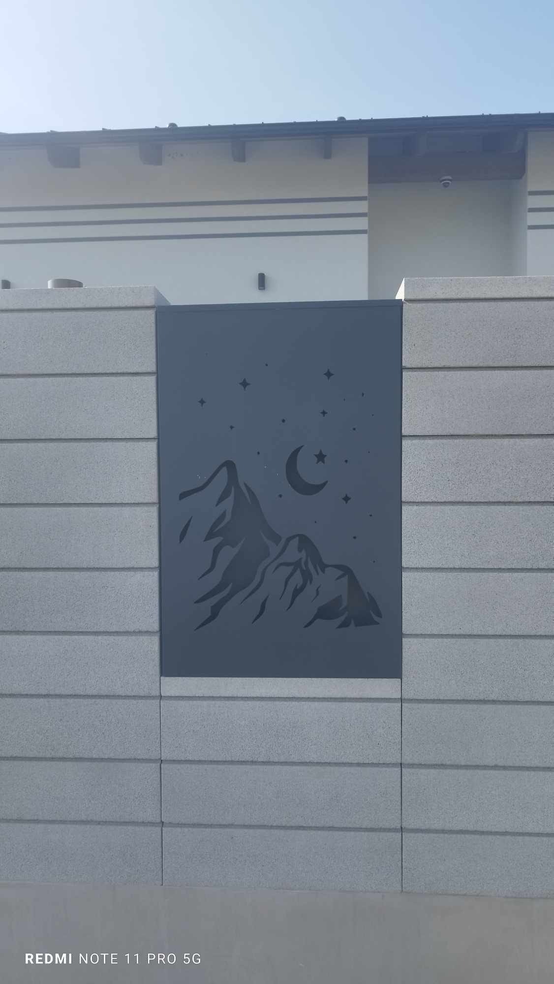 Dekoracyjny panel metalowy z motywem górskim i gwiazd umieszczony w betonowym ogrodzeniu, dodający unikalnego charakteru posesji.