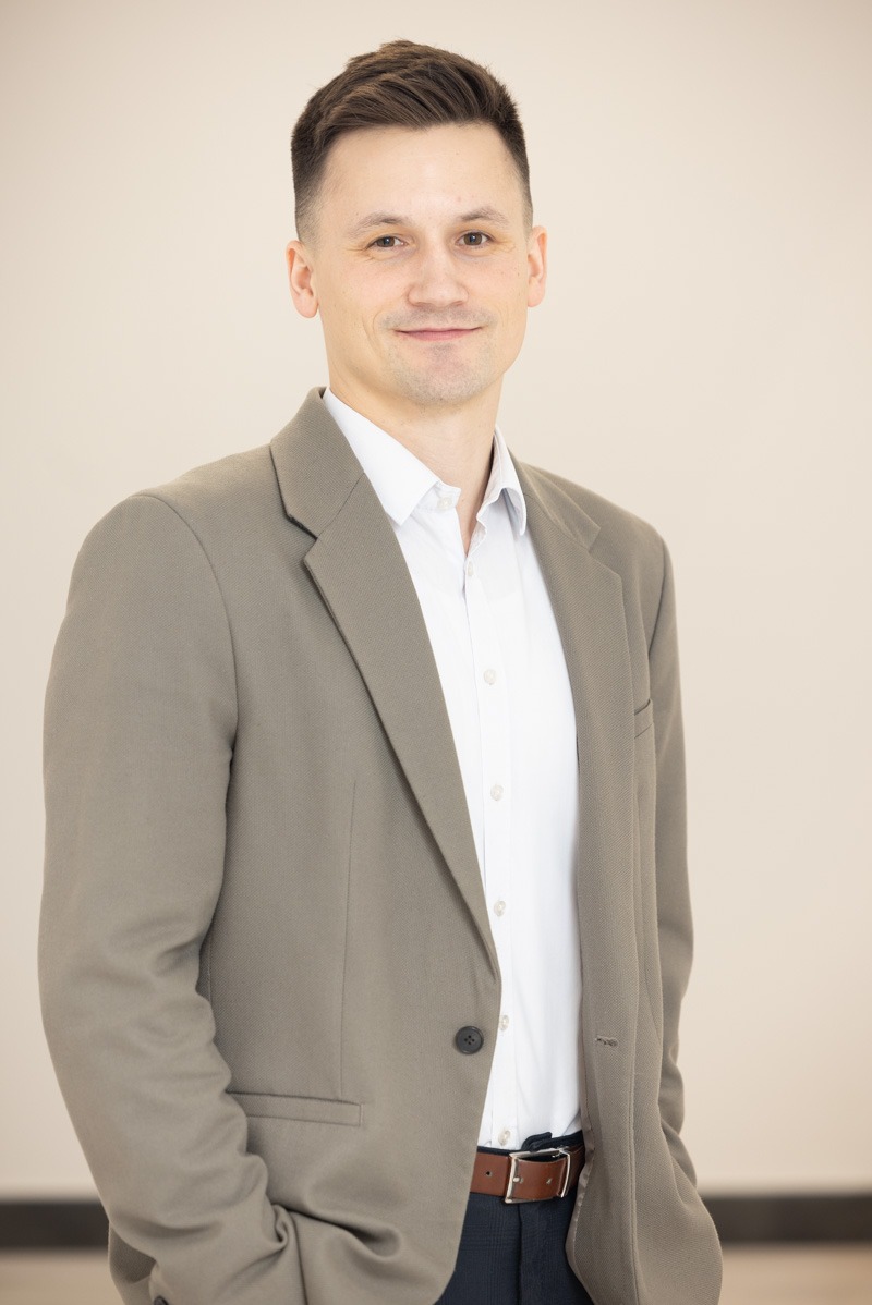 Uśmiechnięty mężczyzna w szarej marynarce i białej koszuli, z brązowym paskiem. Portret biznesowy na jasnym tle, sugerujący profesjonalizm i zaufanie.