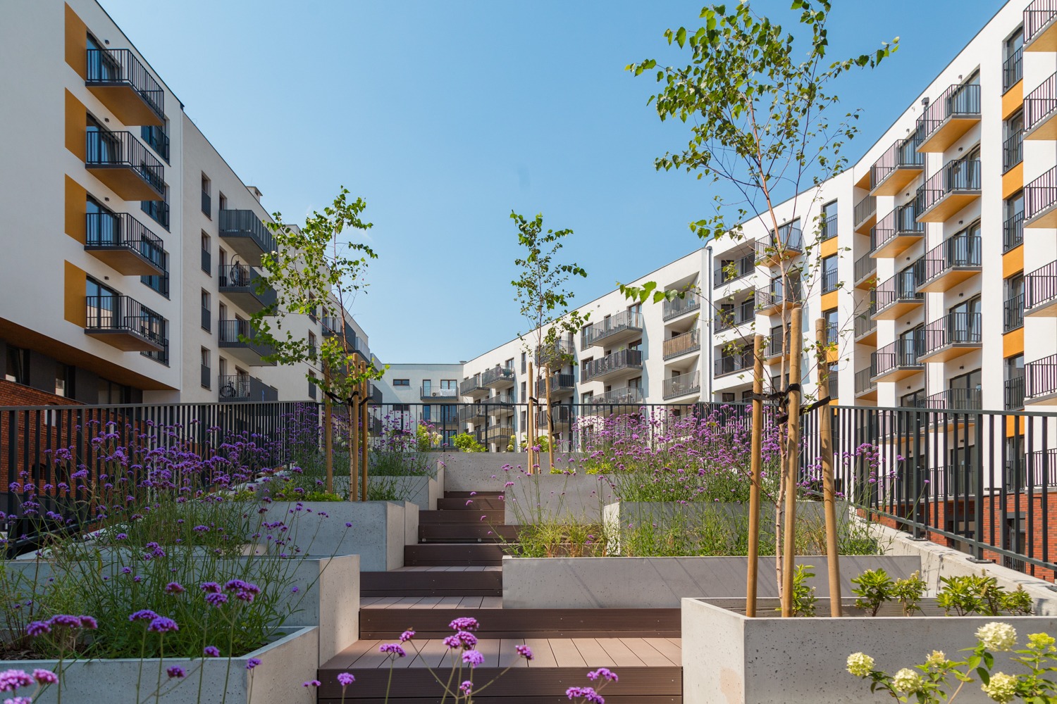 Nowoczesne osiedle z balkonami, schodami i zielenią. Widok na budynki mieszkalne z elementami architektury krajobrazu. Słoneczny dzień, błękitne niebo.