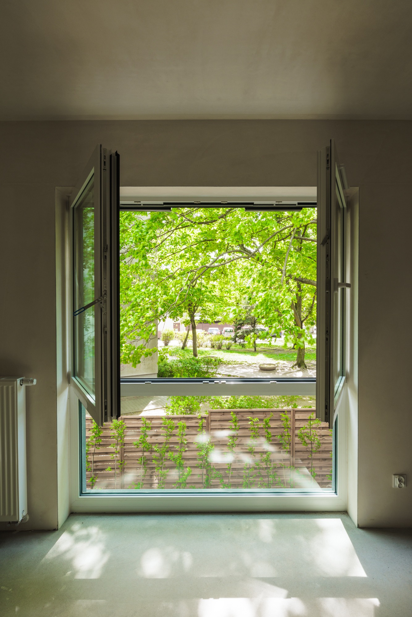 Otwarty balkonowy okno z widokiem na zieleń drzew, widoczny fragment grzejnika i gniazdko elektryczne. Jasne wnętrze, betonowa podłoga w surowym stanie.