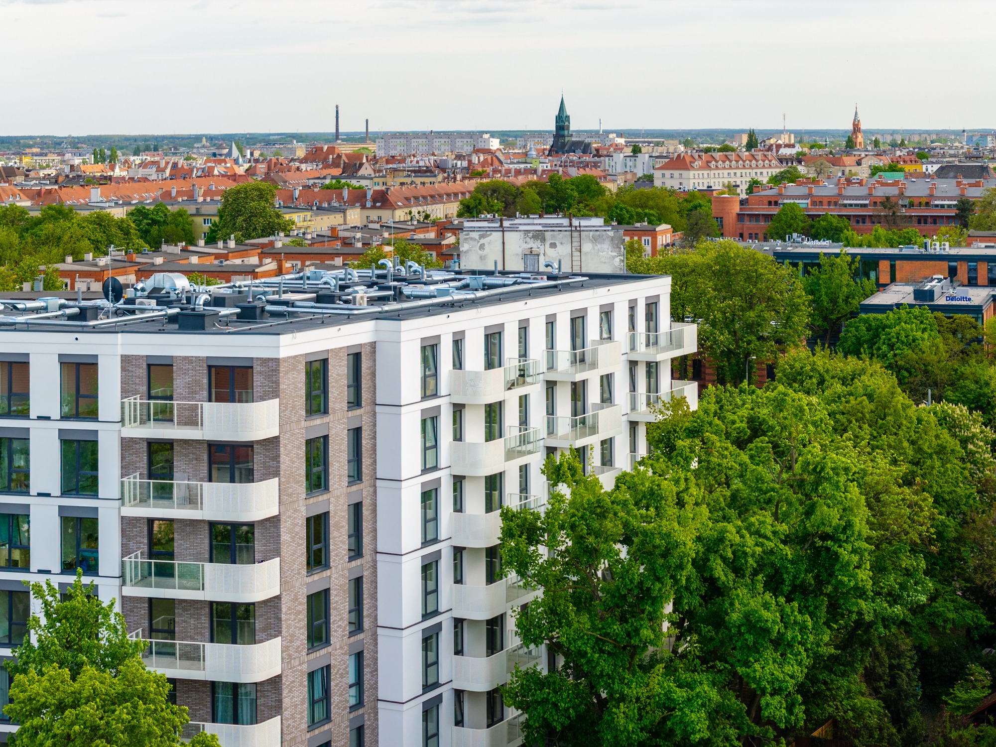 Nowoczesny apartamentowiec z balkonami, widok z góry na panoramę miasta z zielenią. Jasna elewacja z elementami cegły. Dzień, dobre oświetlenie.