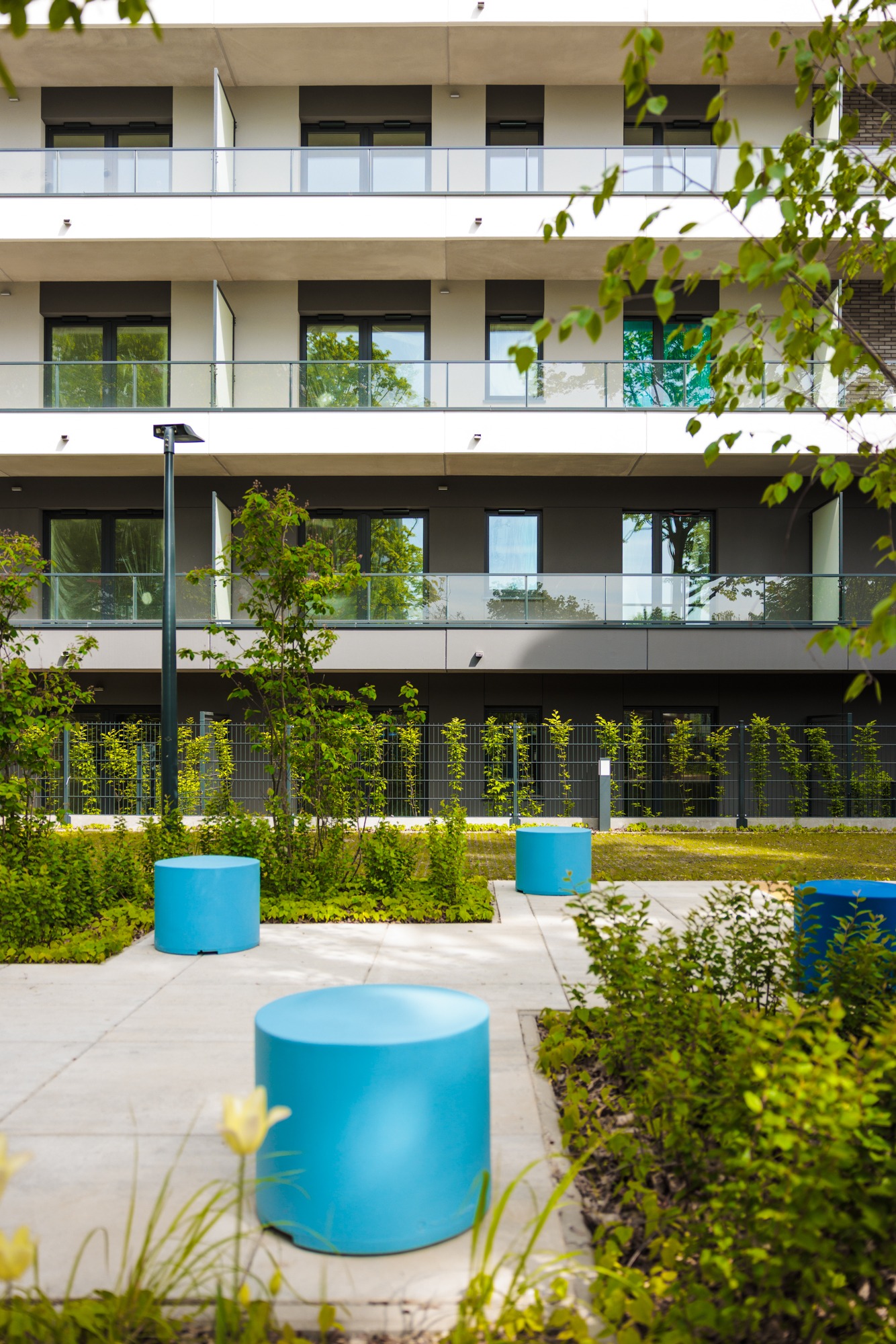 Nowoczesny apartamentowiec z balkonami i zielonym dziedzińcem z niebieskimi siedziskami, w tle widoczna zieleń i ogrodzenie.