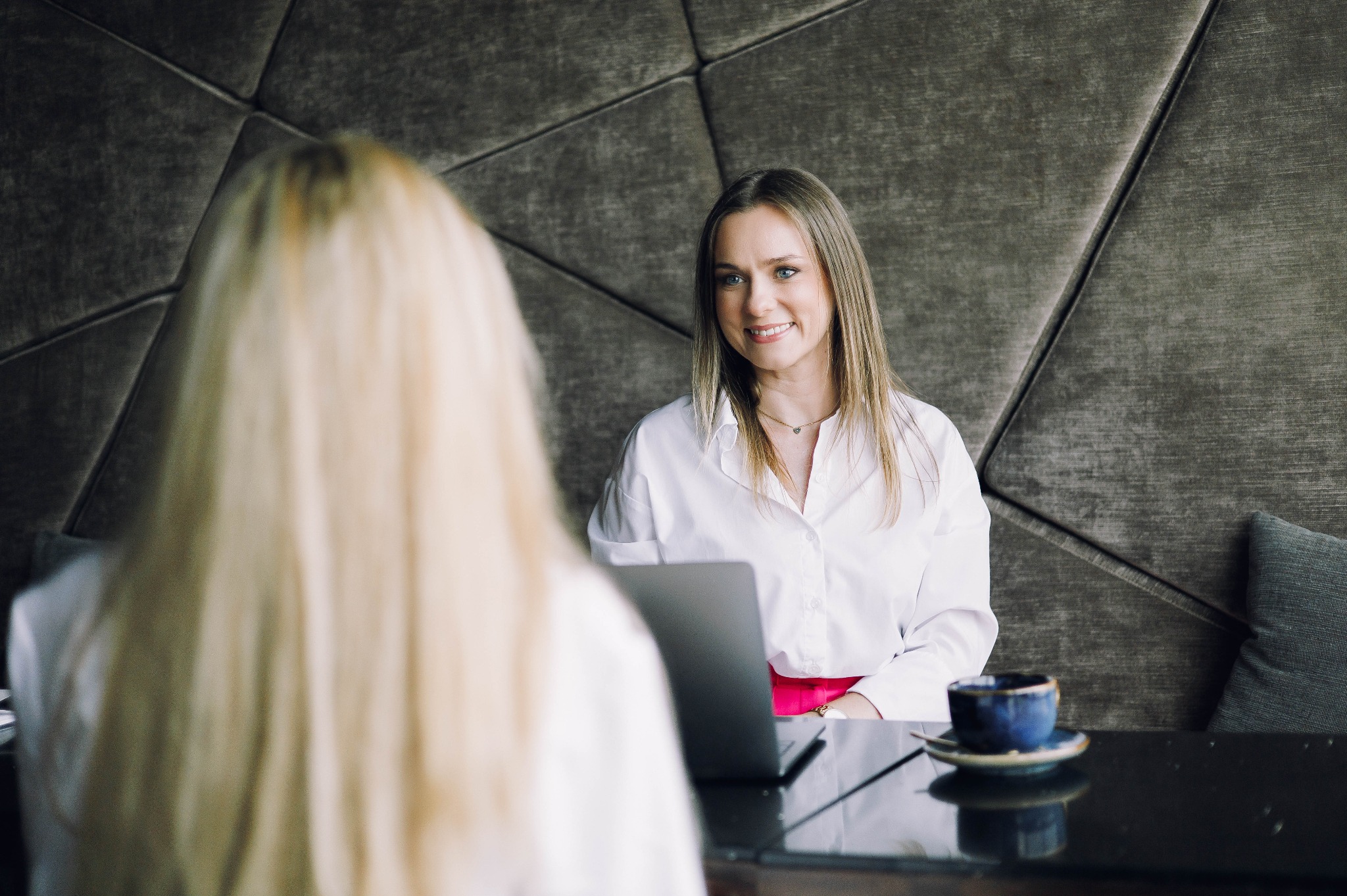 Kobieta w białej koszuli uśmiecha się, siedząc przy stole z laptopem i filiżanką kawy. W tle geometryczna ściana. Rozmowa biznesowa, konsultacja.