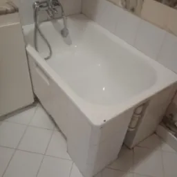 Biała wanna z baterią i słuchawką prysznicową w łazience z białymi kafelkami. Widok z góry na czystą wannę i fragment podłogi z jasnych płytek.